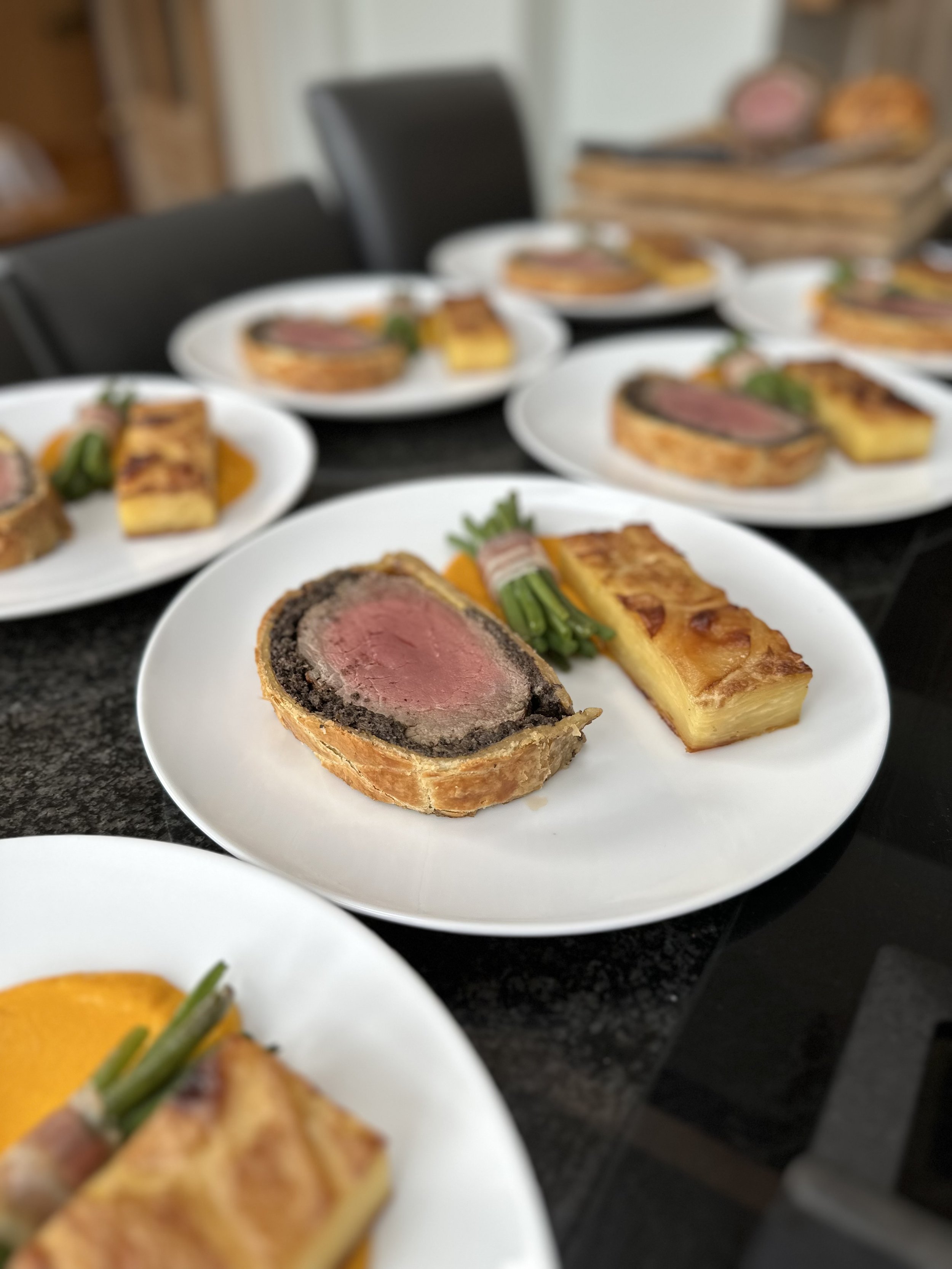 Multiple plates of medium-rare beef Wellington with golden pastry crust and green garnish, served on a black table.