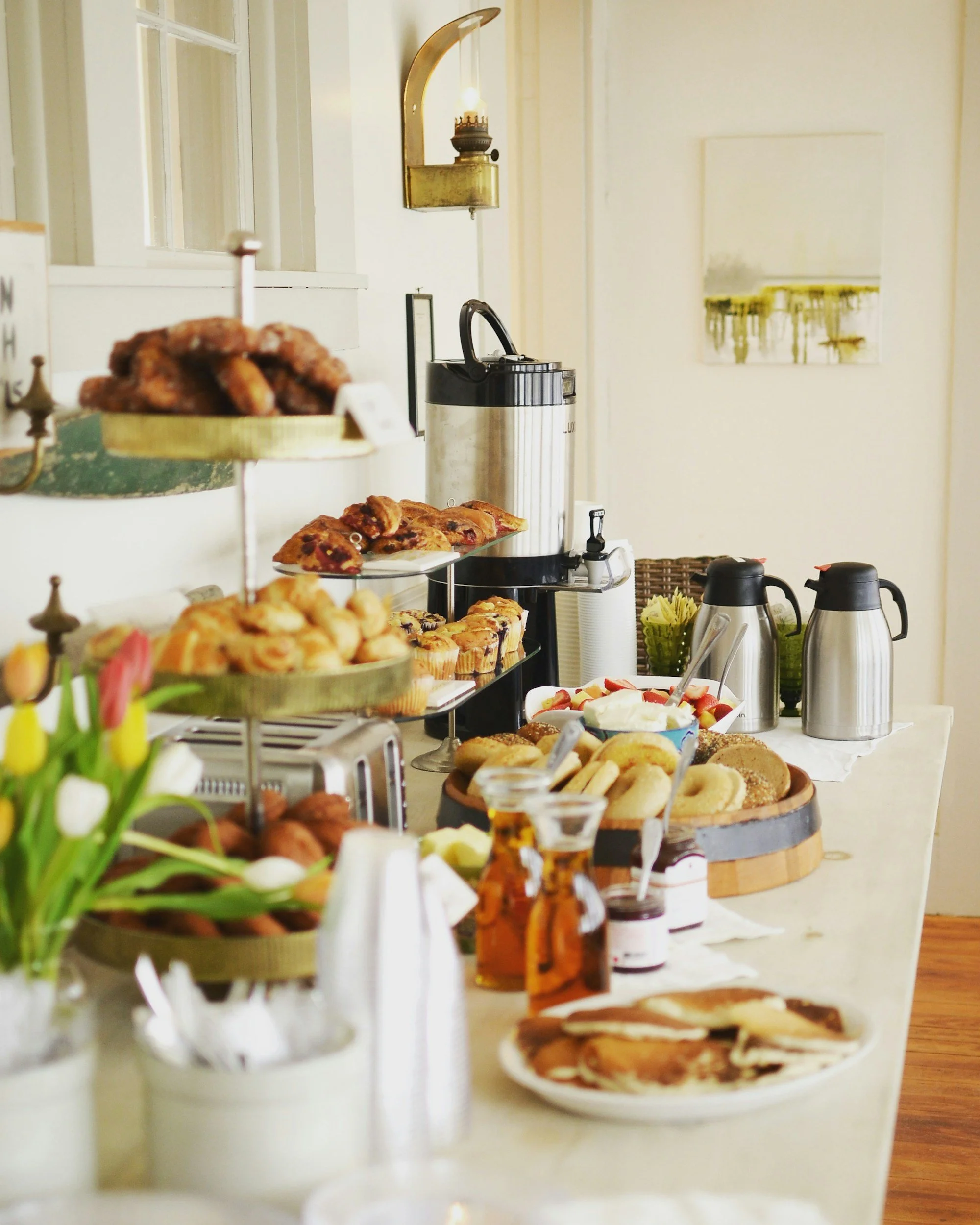 A buffet table filled with assorted pastries, muffins, and cupcakes with a vase of white tulips in the foreground, in a bright room with white walls and a window.