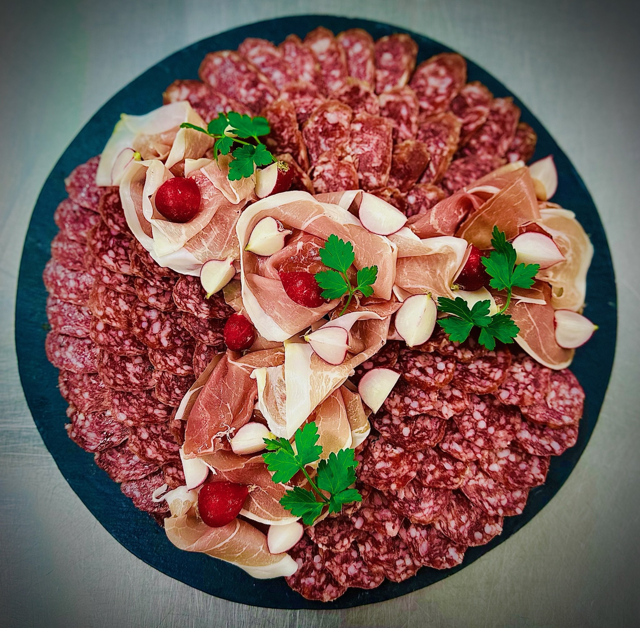 A platter of thinly sliced cured meats, including salami, prosciutto, and other cold cuts, garnished with fresh parsley and radishes.