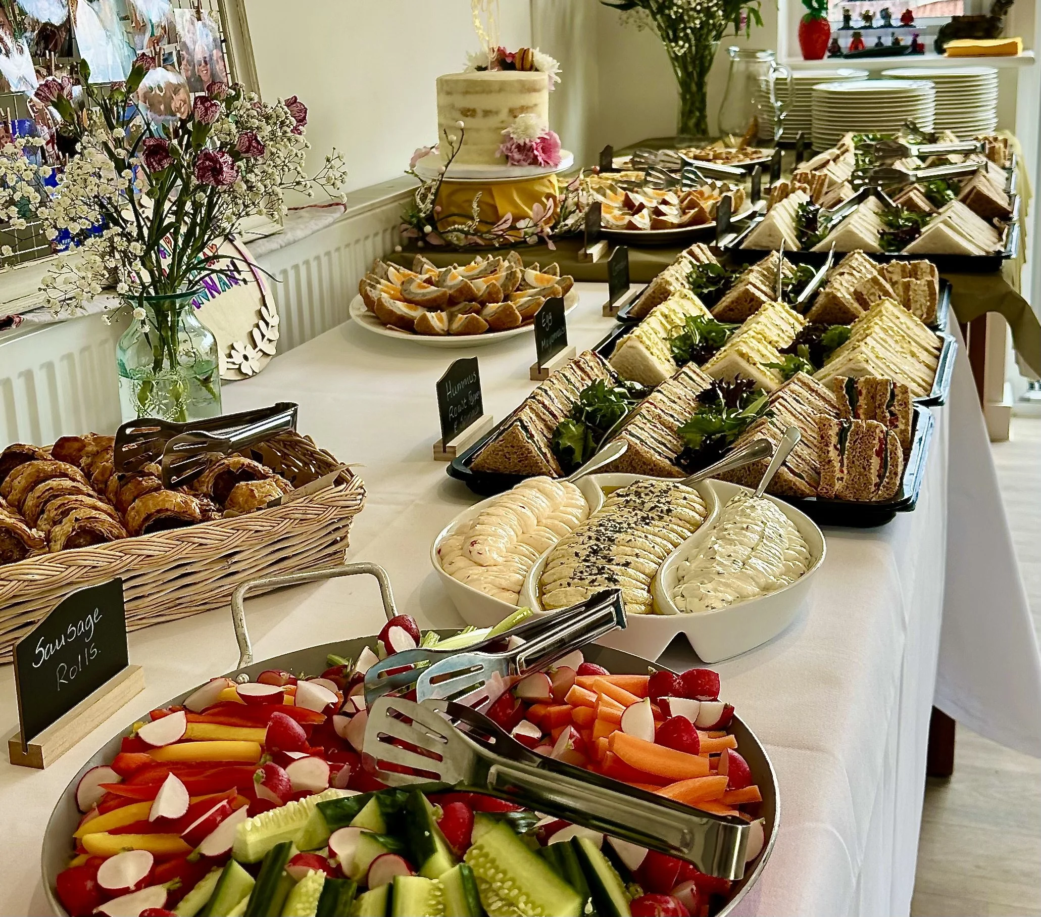 A buffet table with various sandwiches, vegetables, fruits, and desserts, decorated with flowers and a cake in the background.
