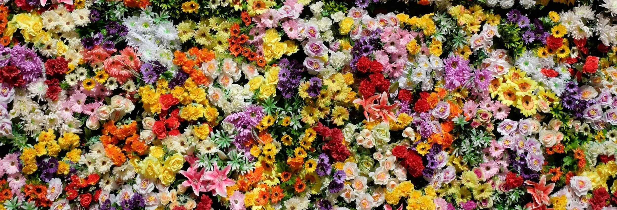 A colourful wall of various fresh flowers, including roses, daisies, and lilies in bright pink, yellow, orange, white, purple, and red hues.