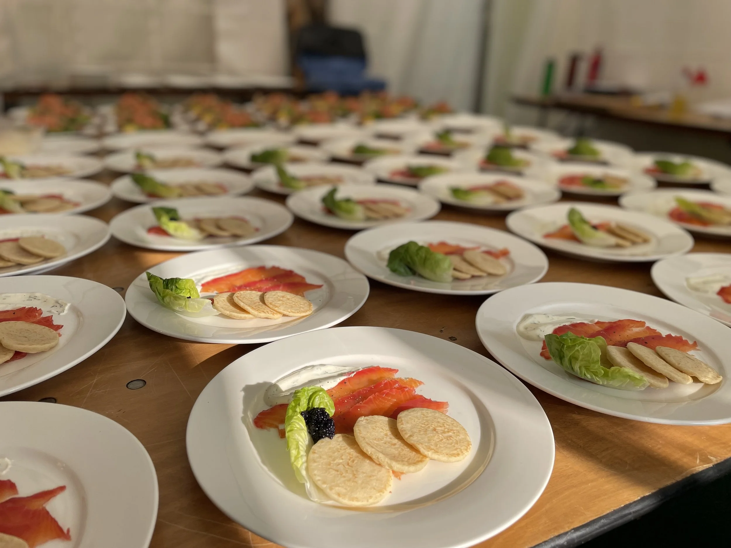 Multiple white plates with smoked salmon, caviar, lettuce, blinis, and cream cheese. Food is arranged on a long table in what appears to be a kitchen or catering setting.