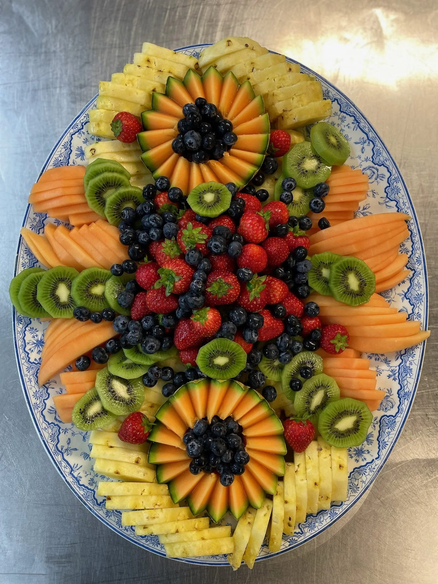A decorative platter of assorted sliced fresh fruits including pineapple, cantaloupe, kiwi, strawberries, and blueberries arranged in a colorful pattern.