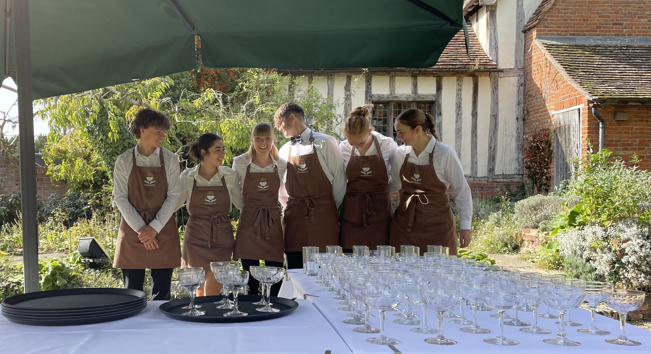 Group of six waitstaff members standing outdoors behind a table set with empty wine glasses, wearing aprons and white shirts, in a garden with old brick and timber buildings in the background.