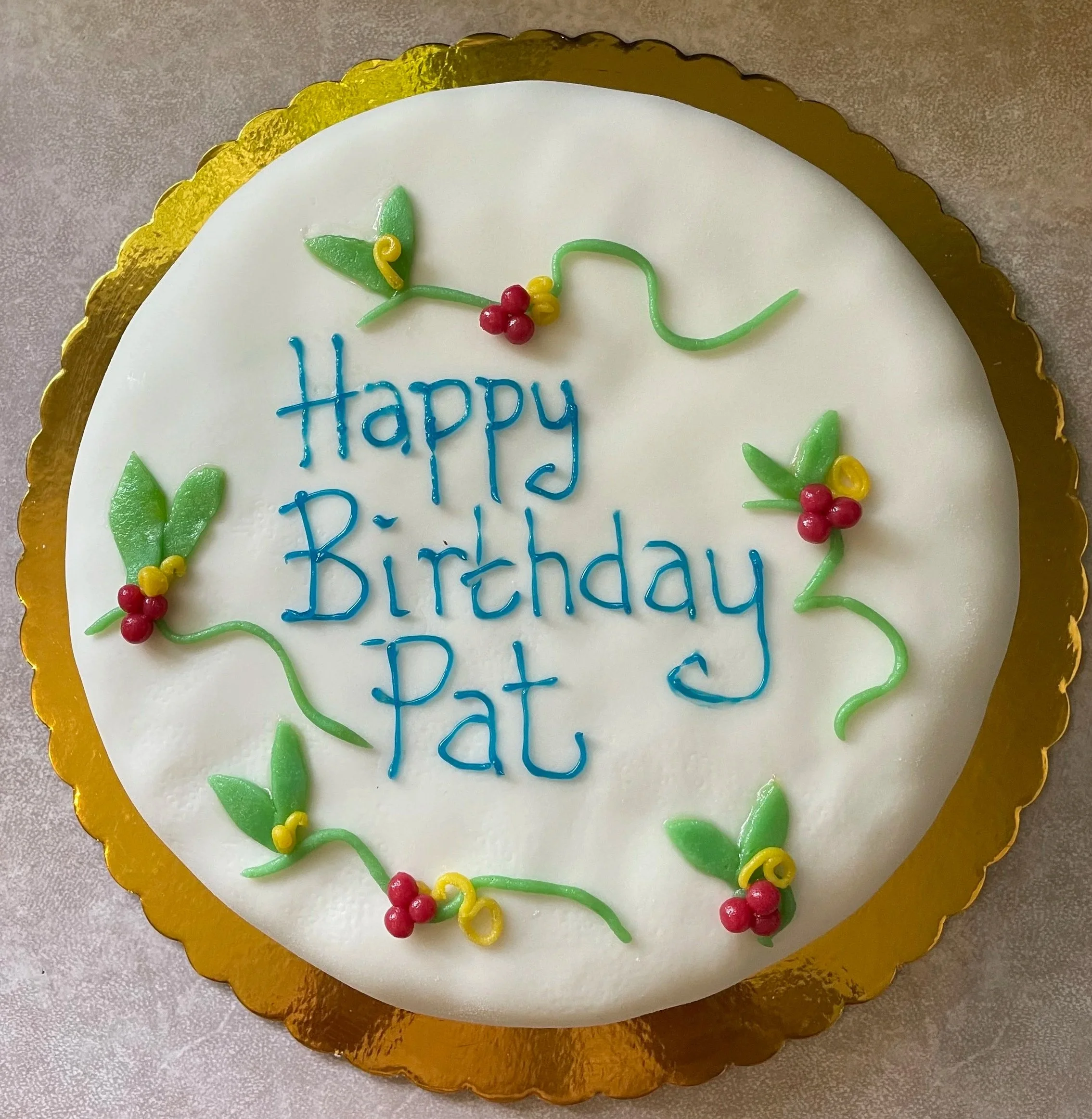 A decorated birthday cake with white icing, colorful icing decorations, and the message 'Happy Birthday' written in blue icing. The cake has a gold scalloped border and is topped with green and red icing decorations resembling leaves and berries.