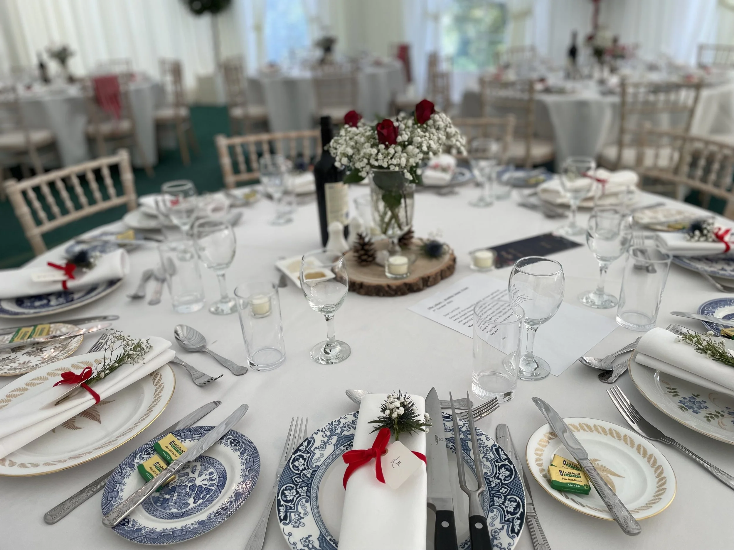 Dining table set for a celebration with white tablecloths, decorated with floral centerpieces, candles, and place settings including blue and white plates, silverware, wine glasses, and napkins tied with red ribbons.