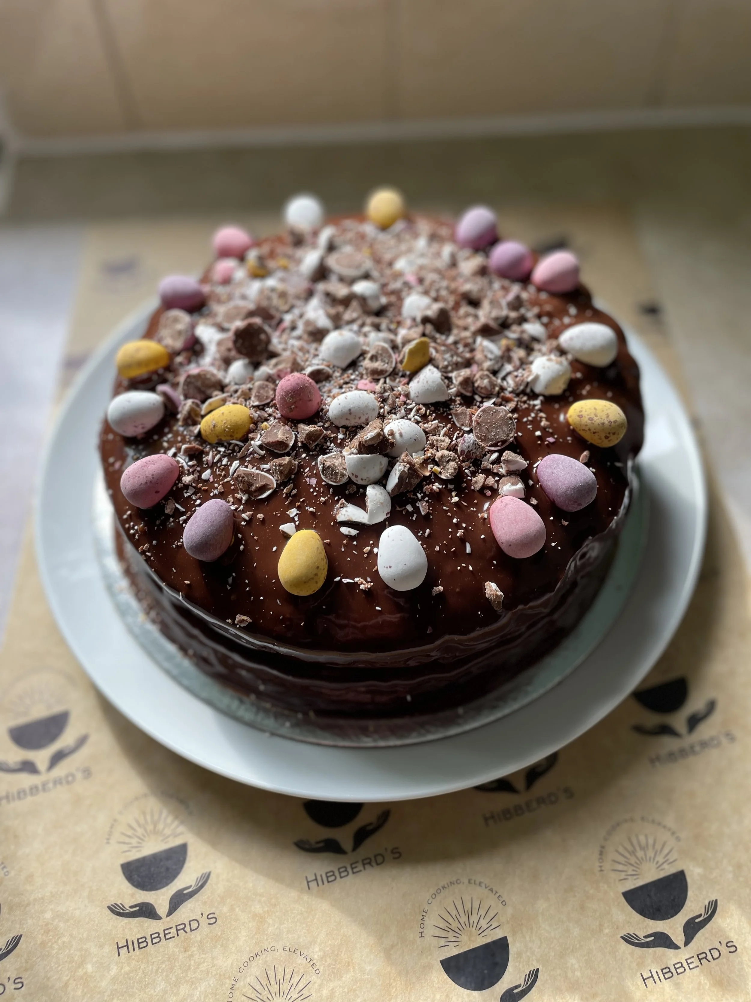 Chocolate cake topped with colourful, crushed chocolates eggs and sprinkles, placed on a white pedestal with Hibberd's branded paper underneath.