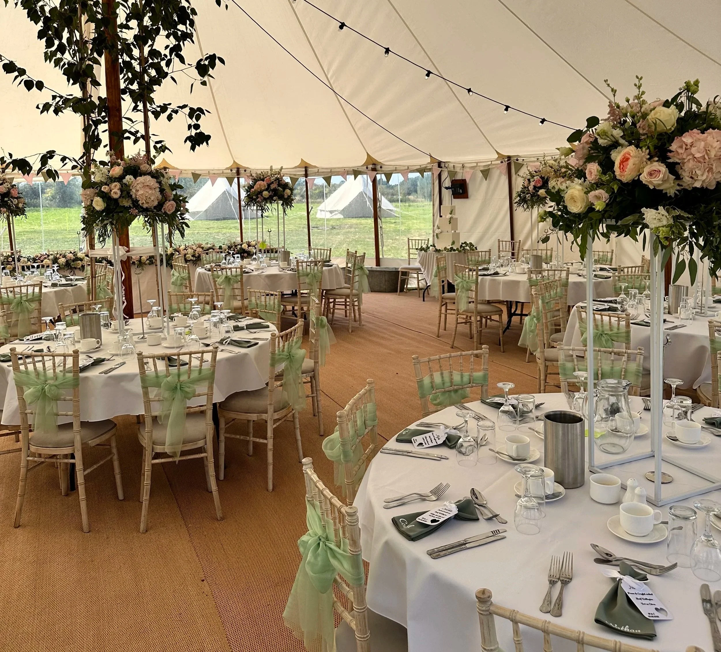 An event tent decorated with floral arrangements and string lights, set up for a celebration or wedding reception, with tables, chairs, and place settings inside.