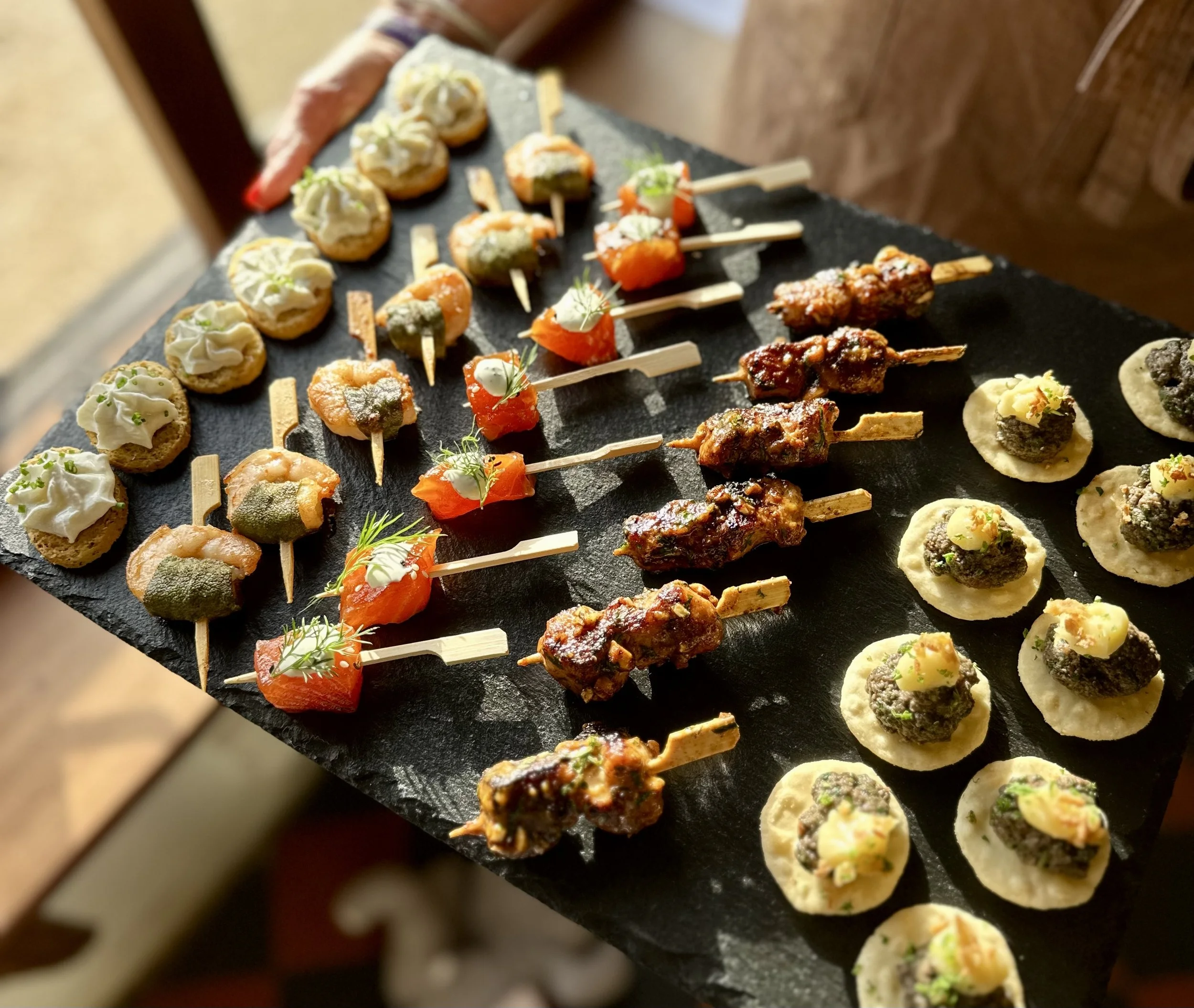 Assorted small canapés on a black serving platter, including skewered meats, seafood, and bites topped with herbs and sauces.