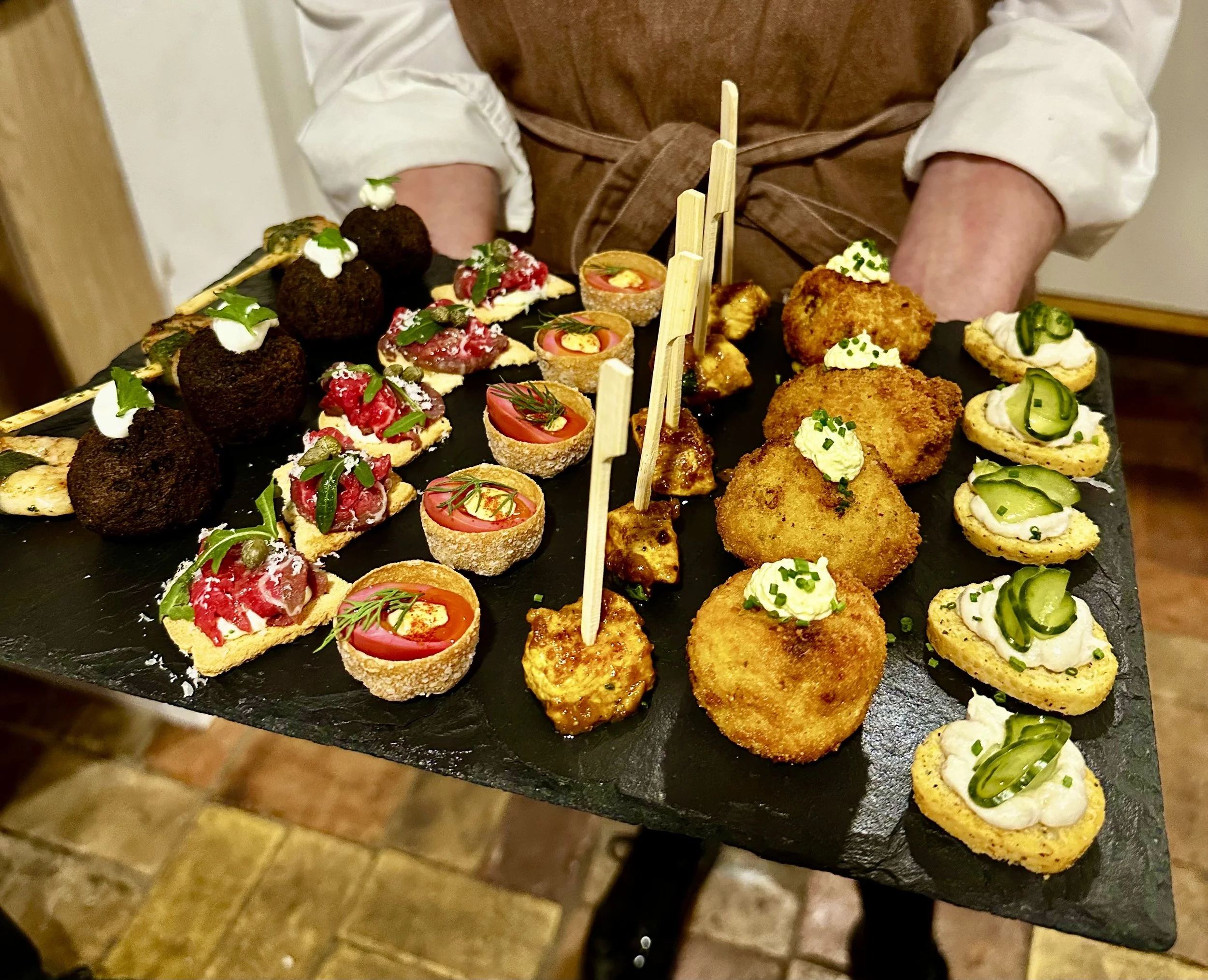 Assorted gourmet canapés on a black slate platter, including meatballs, mini quiches, and crispy stuffed croquettes, garnished with herbs.
