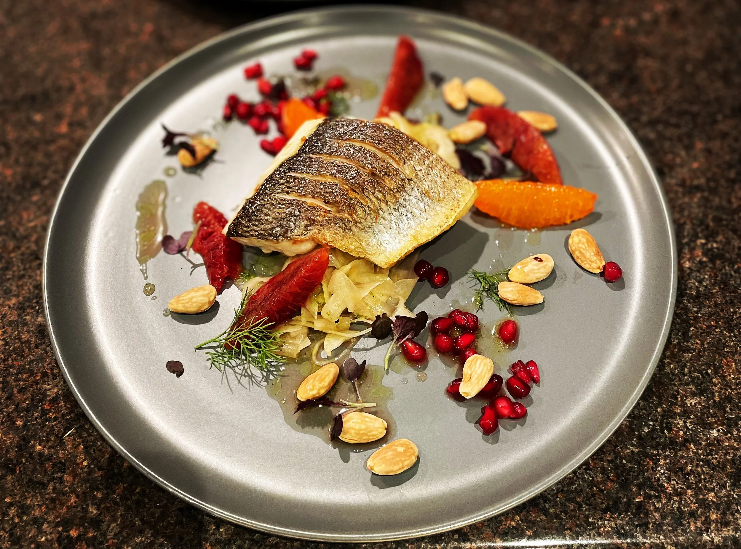 A plated serving of fish on a bed of vegetables, garnished with almonds, pomegranate seeds, and herbs.