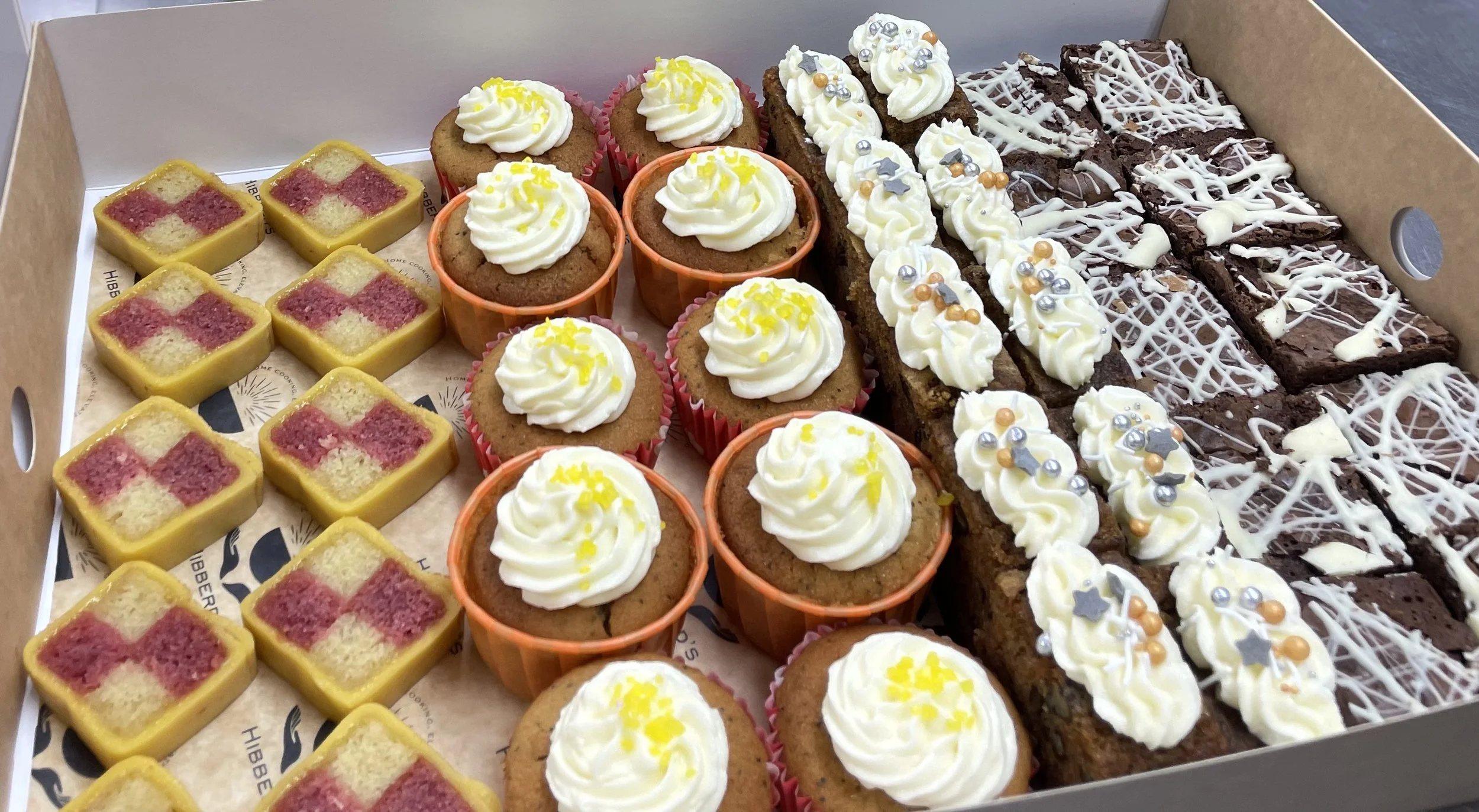 Assorted baked goods including small square cakes with pink and yellow pattern, cupcakes with white frosting and yellow sprinkles, and brownies decorated with white chocolate drizzle and colorful sprinkles inside a cardboard box.
