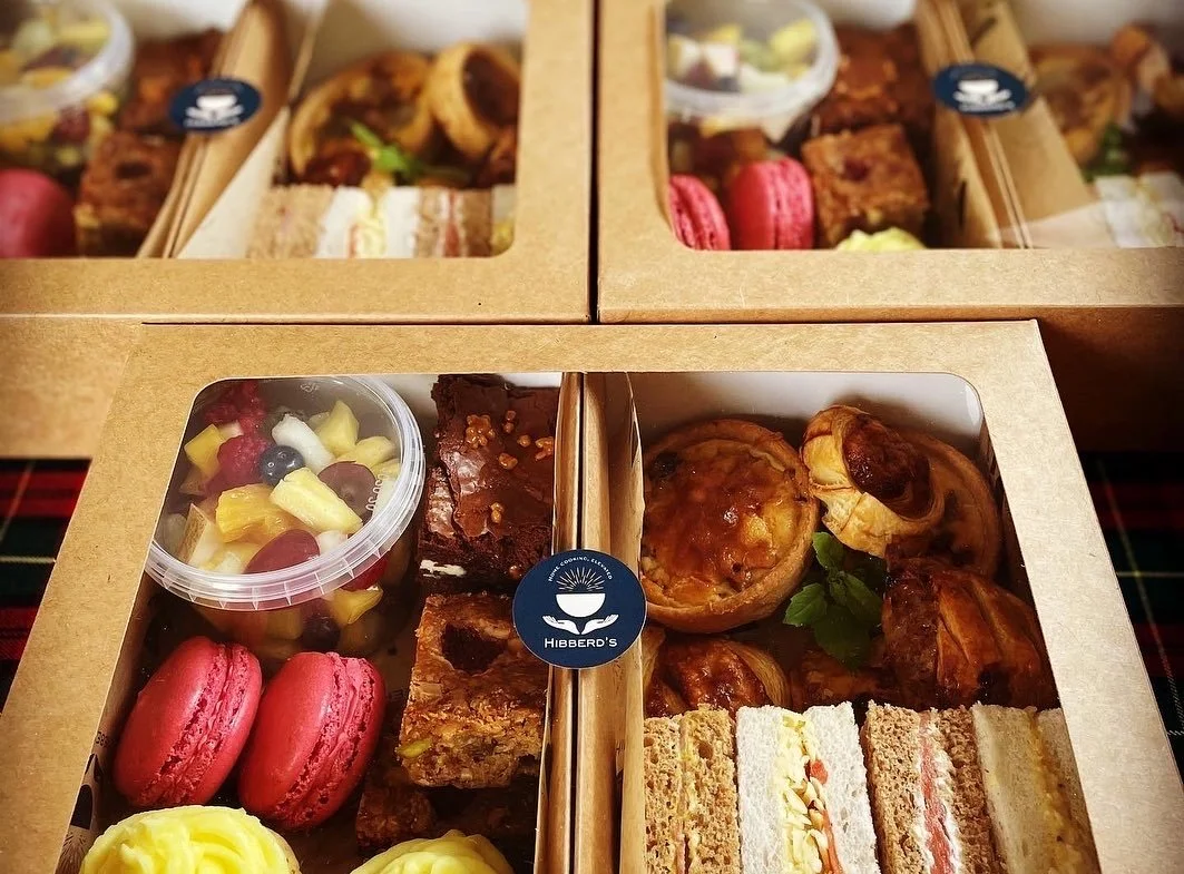 Assorted afternoon tea boxes containing muffins, macarons, fruit, cakes, and sandwiches, labeled 'Hibberd's'.