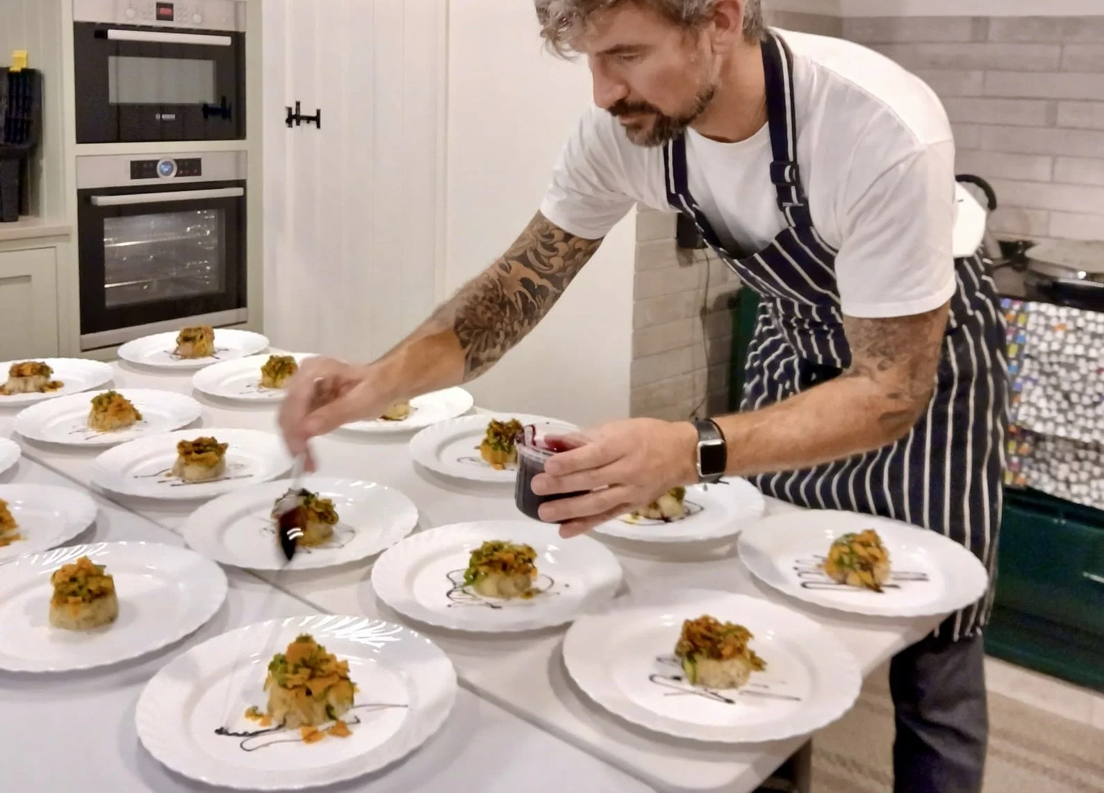 A handsome chef with tattoos on his arms is preparing multiple plates of gourmet food, pouring a sauce over each dish in a kitchen setting.