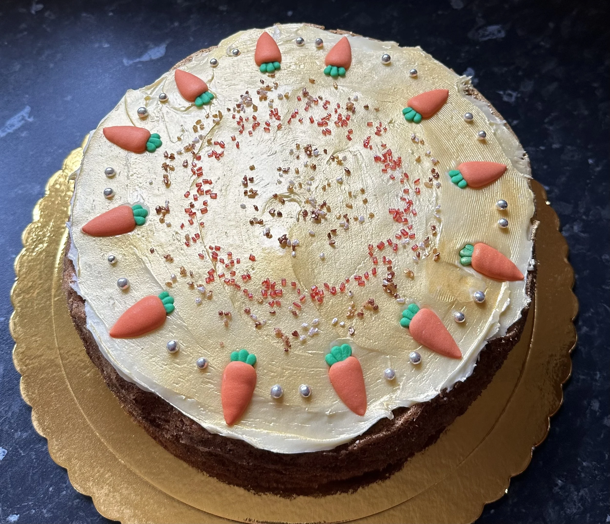 A round birthday cake with gold-colored icing, decorated with orange and green fondant carrots and silver, red, and gold sprinkles, on a black surface.