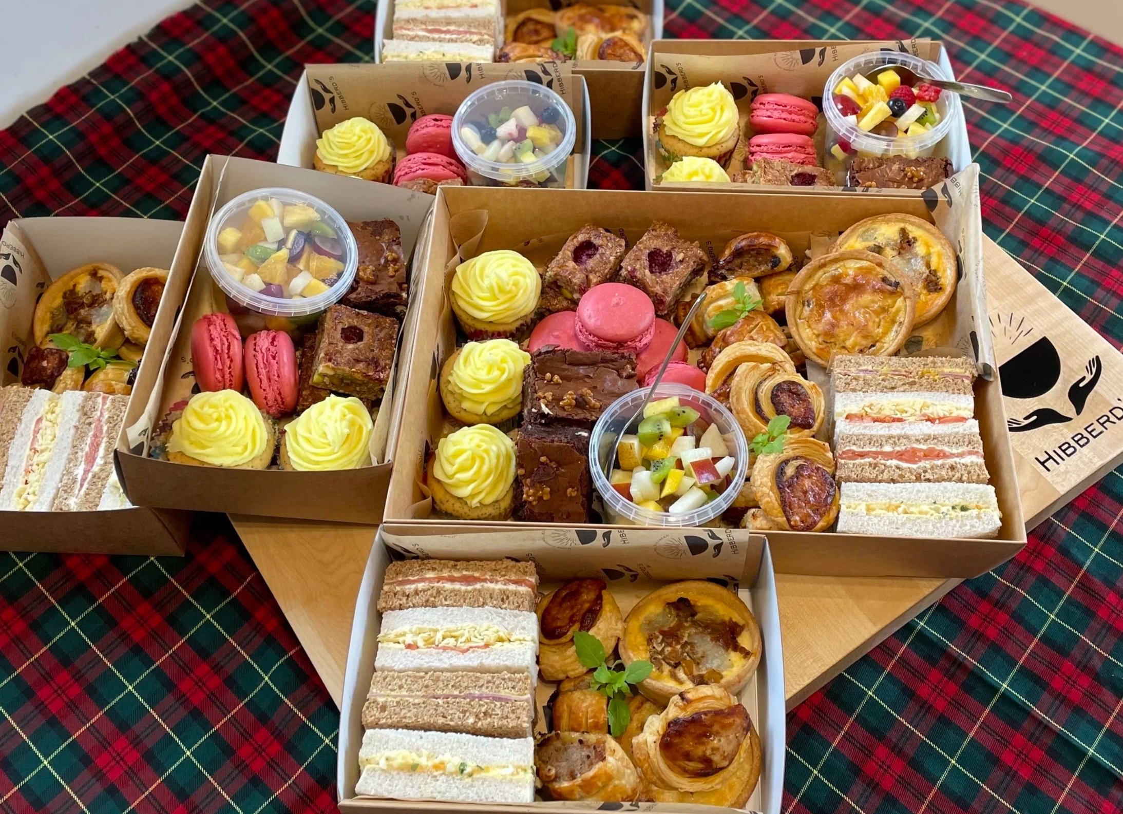 Assorted pastries, sandwiches, macarons, fruit cups, and cupcakes on a holiday plaid tablecloth.