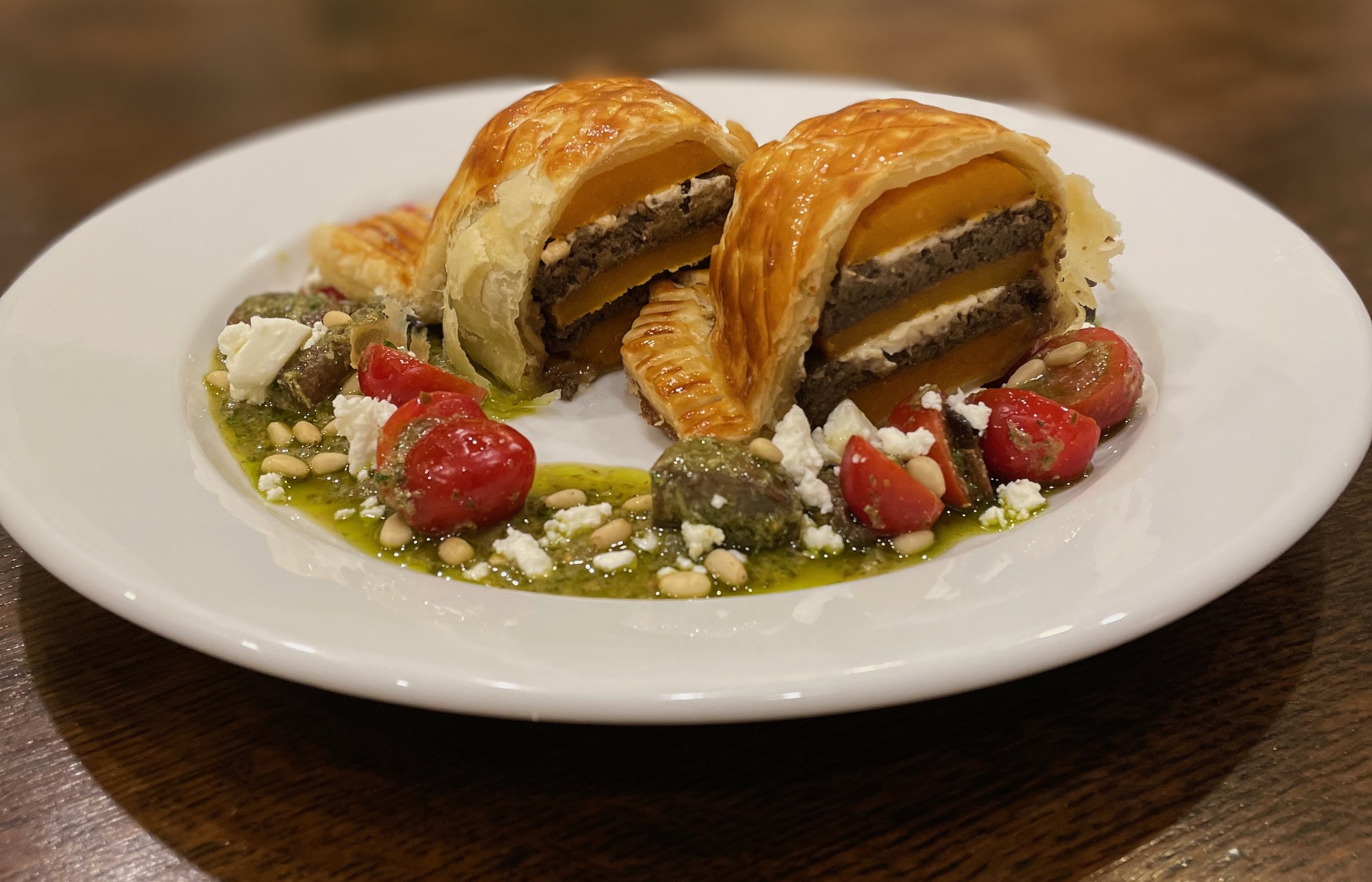 A plate of Vegetarian Wellington with cherry tomatoes, green pesto, pine nuts, and crumbled Feta cheese.