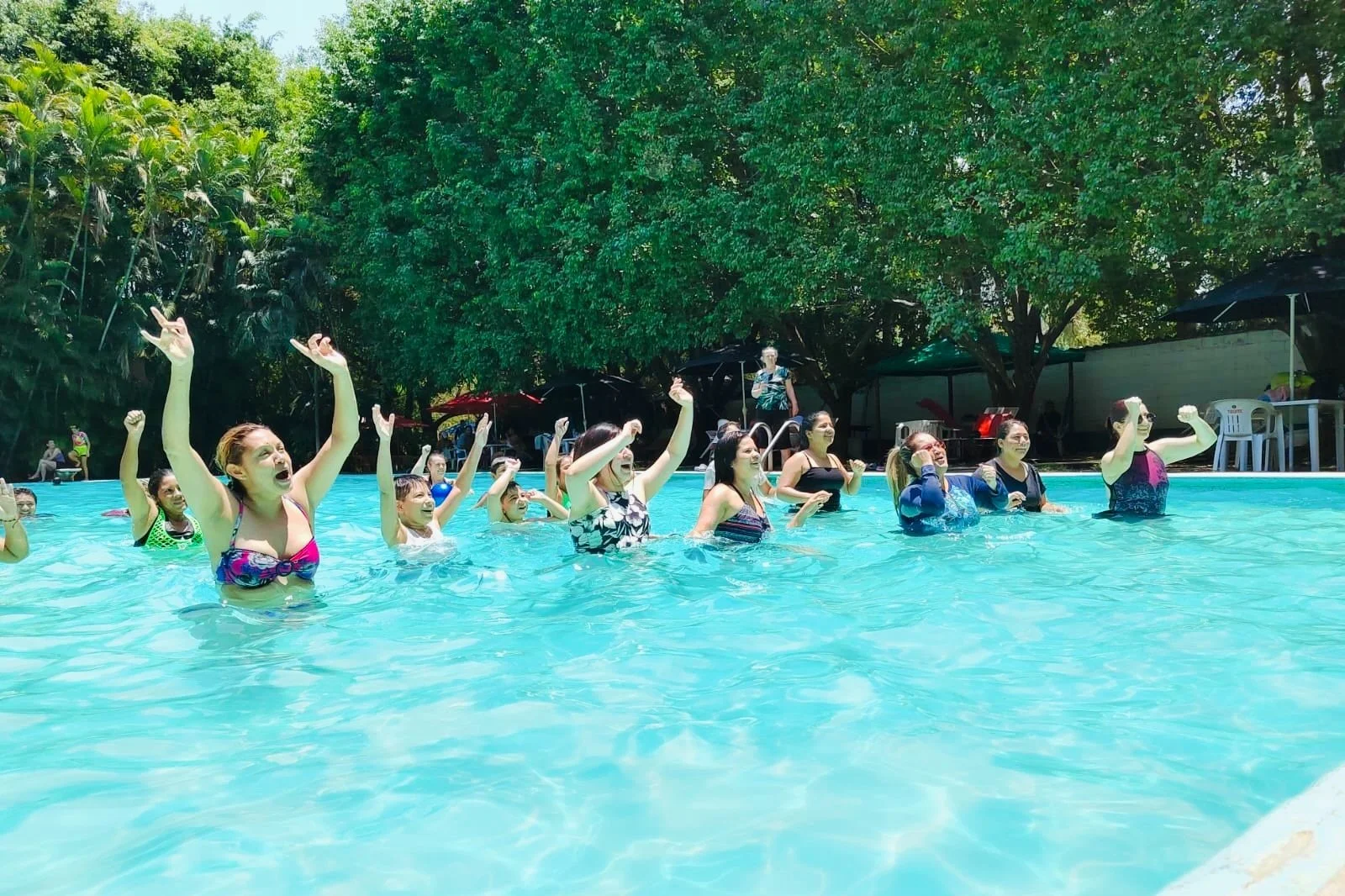 Grupo de mujeres en la alberca, parece que están haciendo una clase de aeróbicos acuáticos, rodeadas de árboles verdes y sombra, con algunos niños y adultos en el fondo.