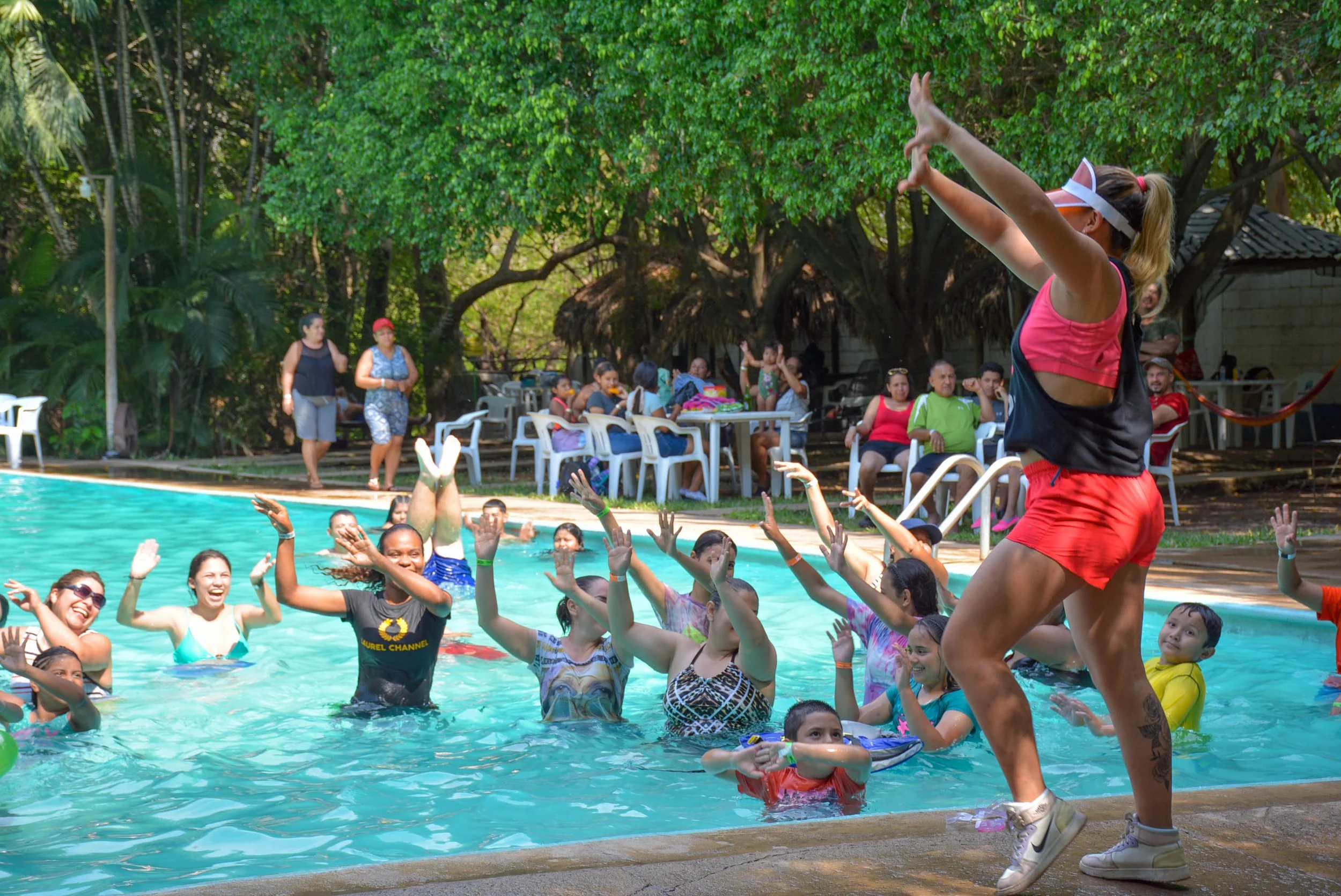 Grupo de personas en una piscina disfrutando de una clase de ejercicio acuático dirigida por una instructora con gafas y visera en un entorno natural con árboles y mesas en el fondo.
