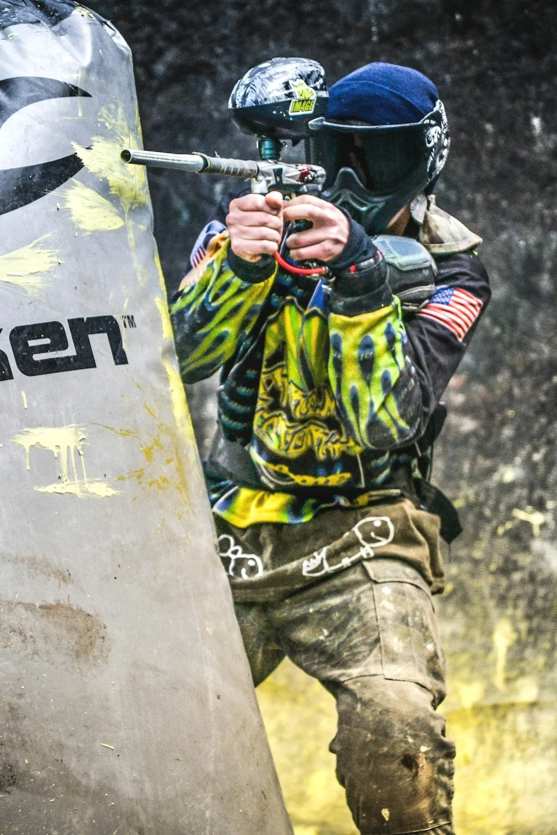 Persona jugando paintball con uniforme, máscara y protecciones, sosteniendo un arma de paintball detrás de un escudo.