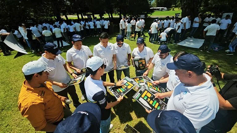 Grupo de personas participando en una actividad al aire libre, revisando pizarras de colores, en un parque con árboles y césped.