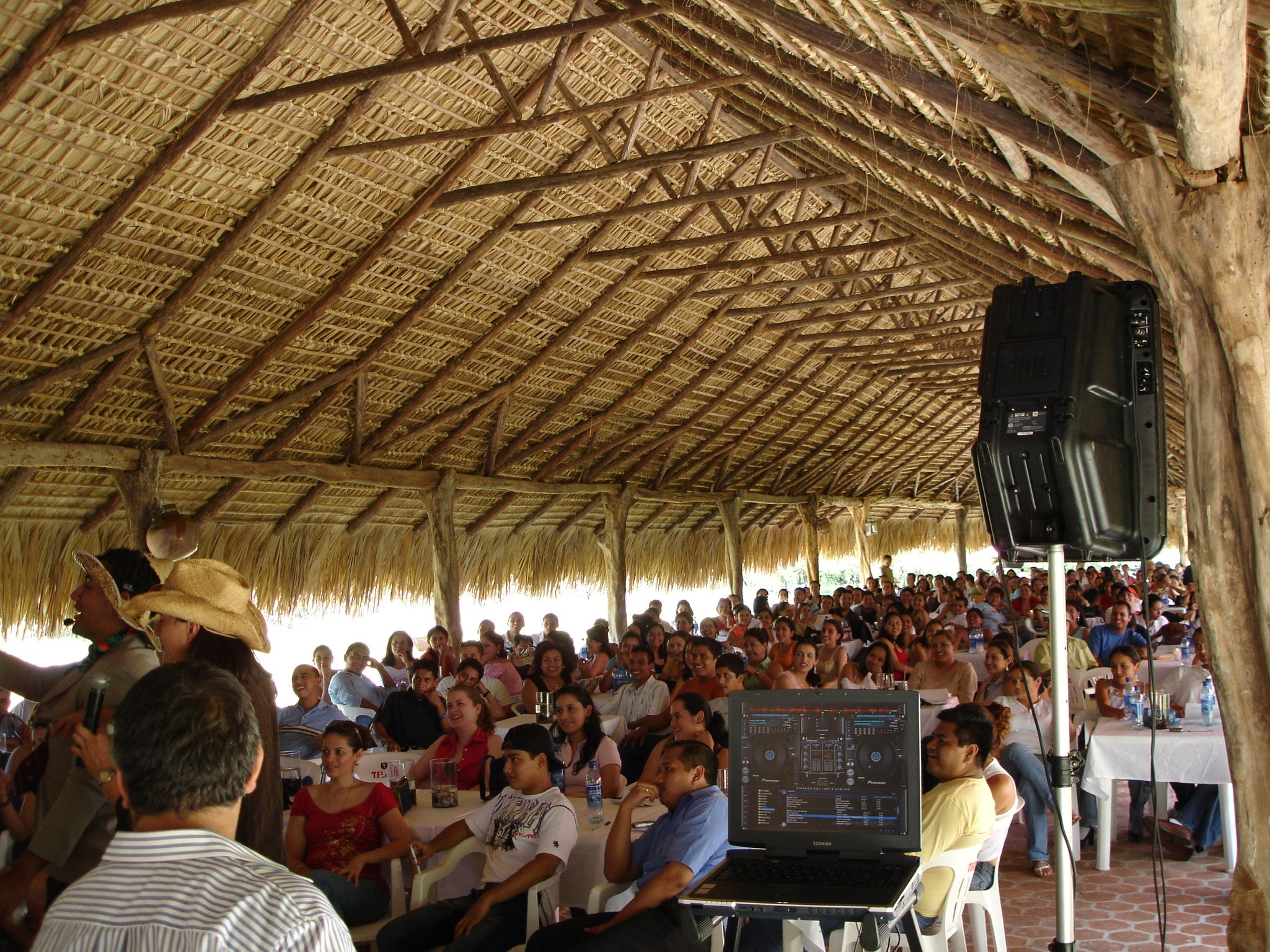 Conferencia o evento social en un lugar abierto con techo de hojas de palma y muchas personas sentadas, algunos con sombreros, y un equipo de sonido y DJ en primer plano.
