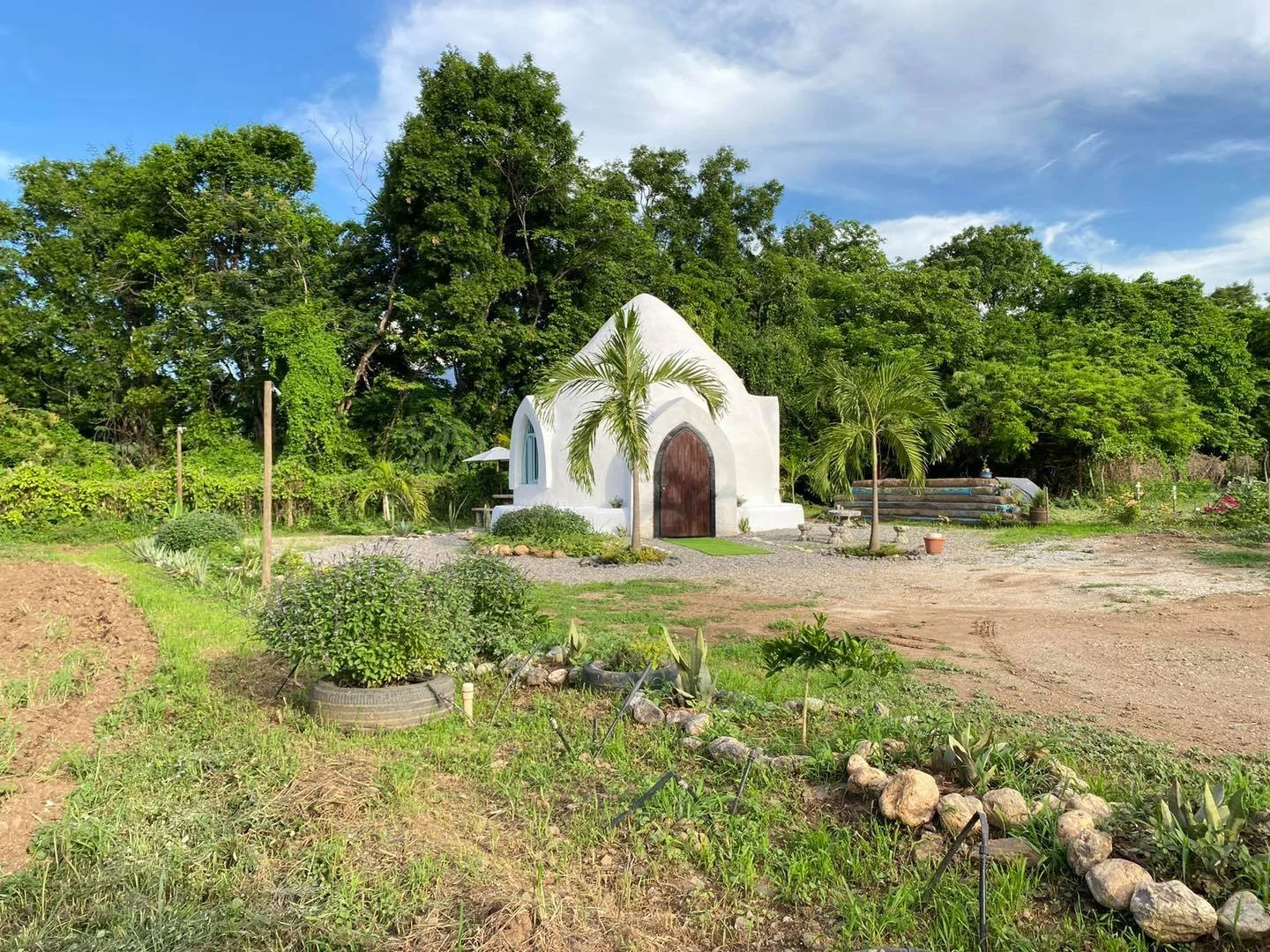 Pequeña capilla blanca con puerta de madera, rodeada de plantas y árboles, en un entorno natural, con cielo parcialmente nublado.