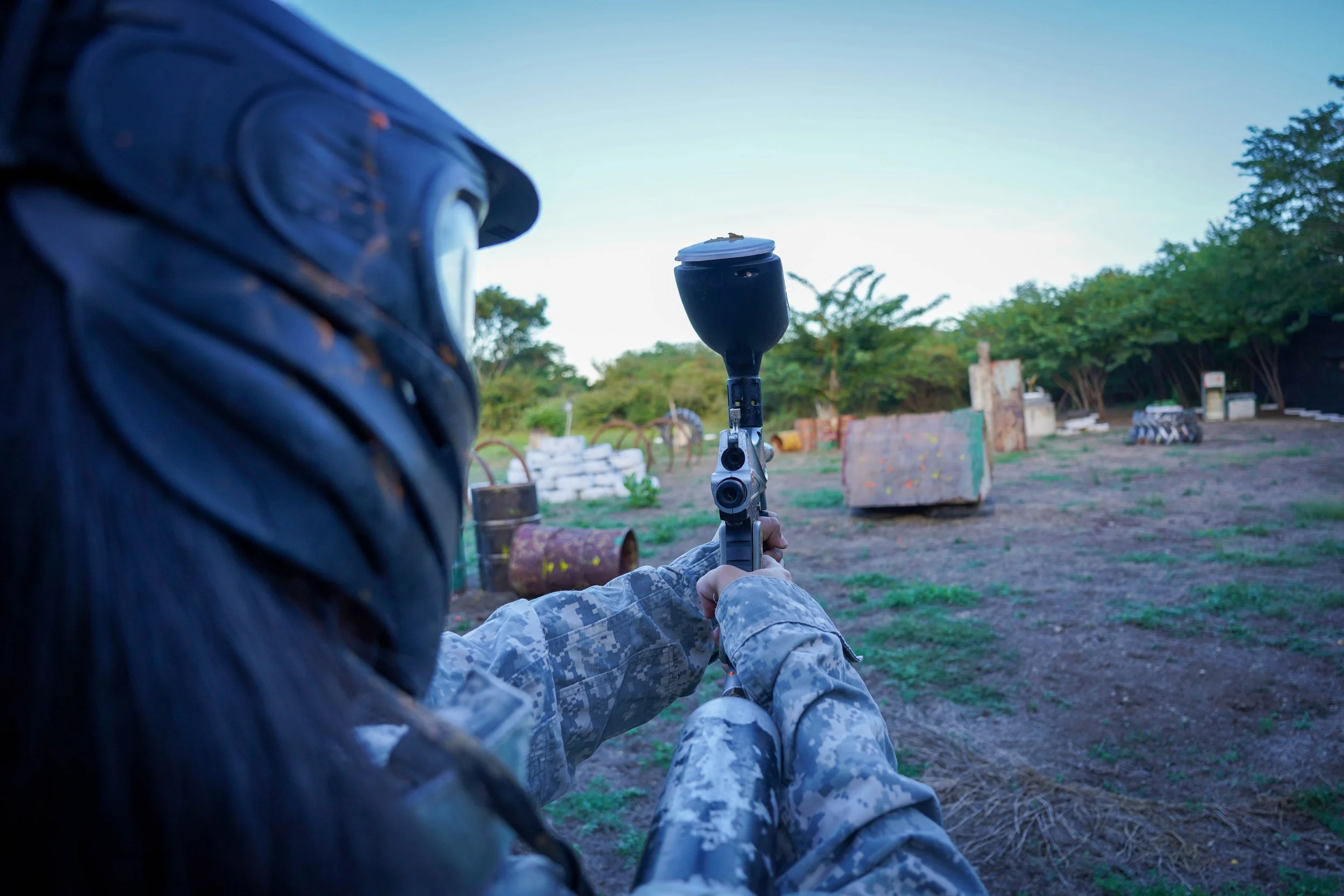 Persona con uniforme de combate en un campo de entrenamiento, apuntando con un rifle de paintball o láser hacia objetos en el suelo.