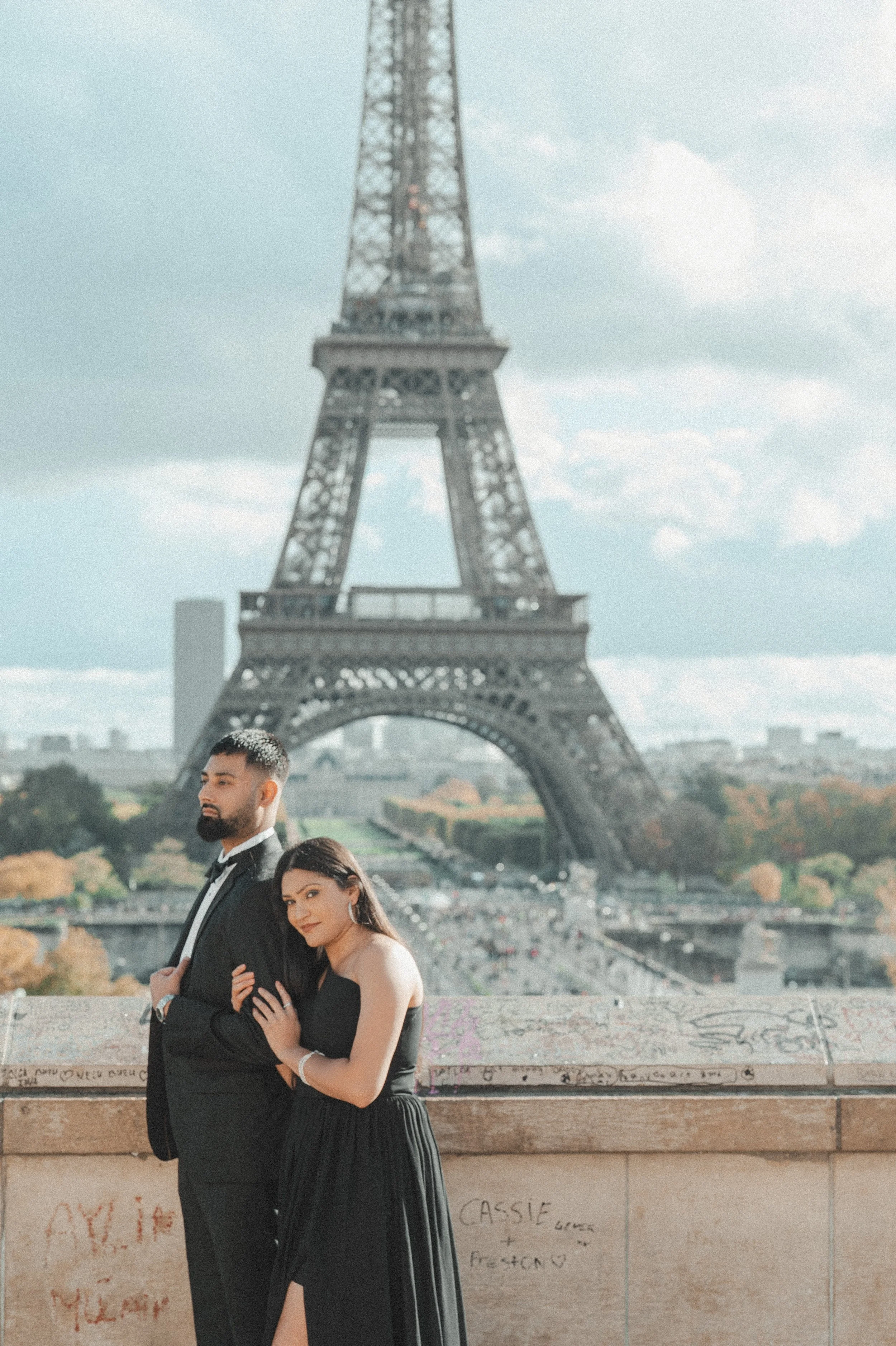 A couple dressed in formal attire standing on a balcony with the Eiffel Tower in the background.
