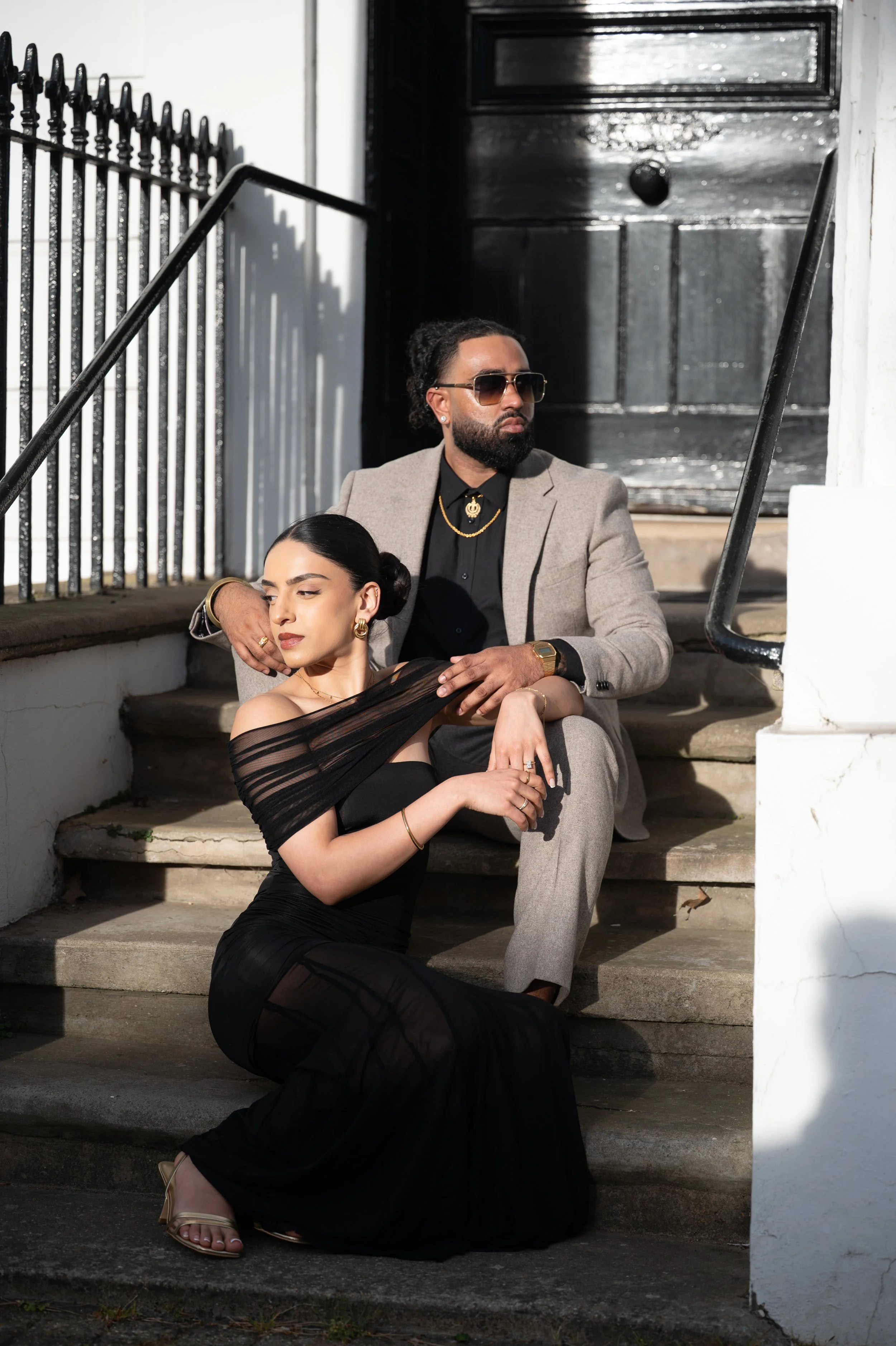 A man and woman dressed in formal attire sitting on stairs outside a white building with black door and railings. The woman is in a black dress, and the man is in a grey suit with sunglasses, jewelry, and a watch.