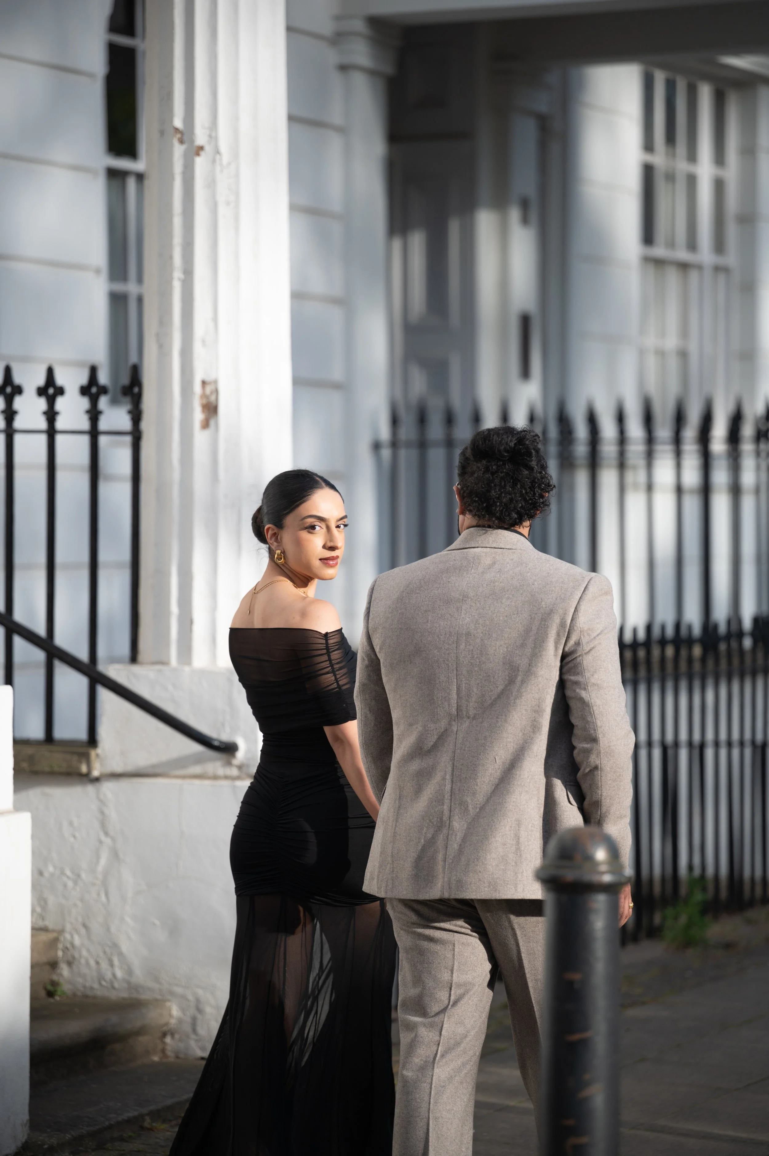 A woman in a black strapless off-shoulder dress and gold earrings stands next to a man in a gray suit on a city street at night. They are near a white building with black iron fencing, and there is a black bollard in the foreground.