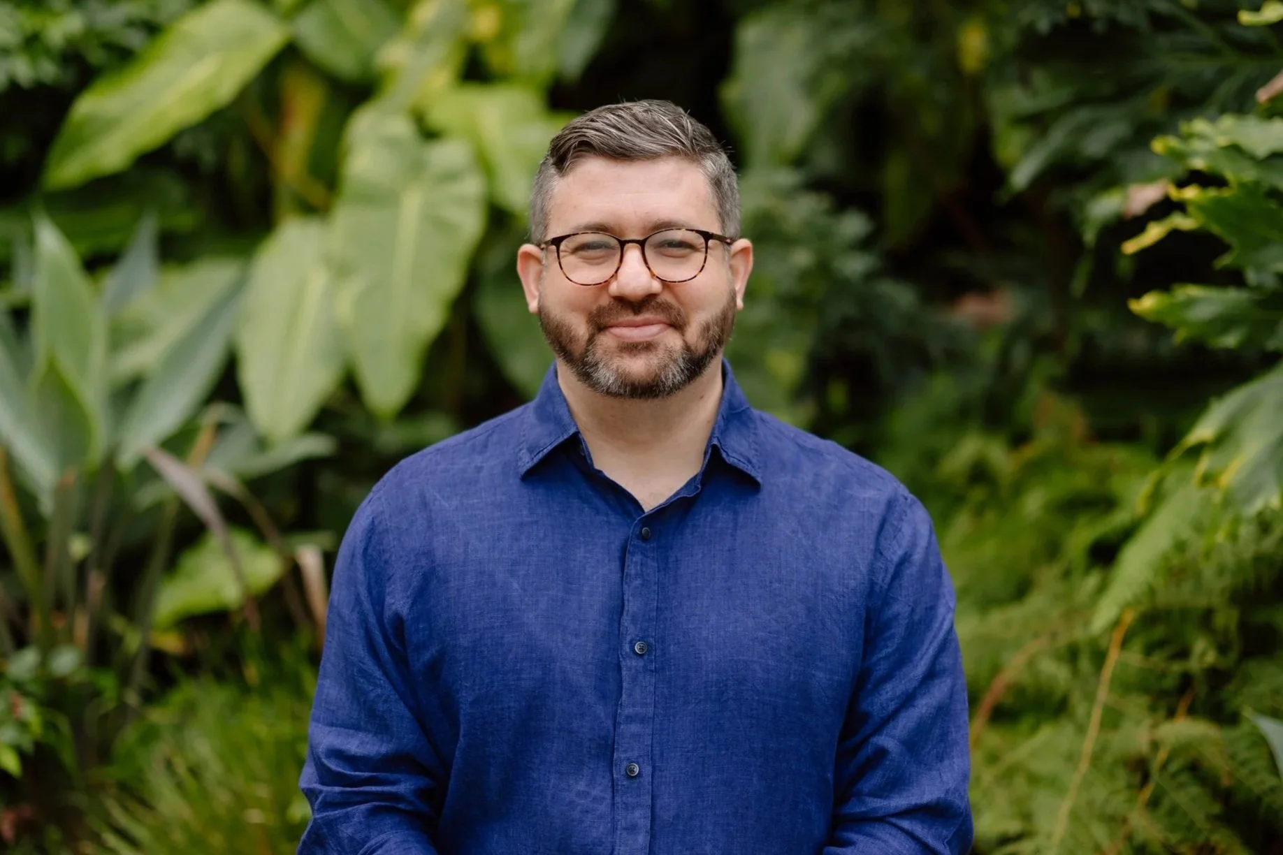 A young man with glasses, a beard, and dark hair, wearing a navy sweater and jeans, standing outdoors against a brick wall, smiling at the camera.