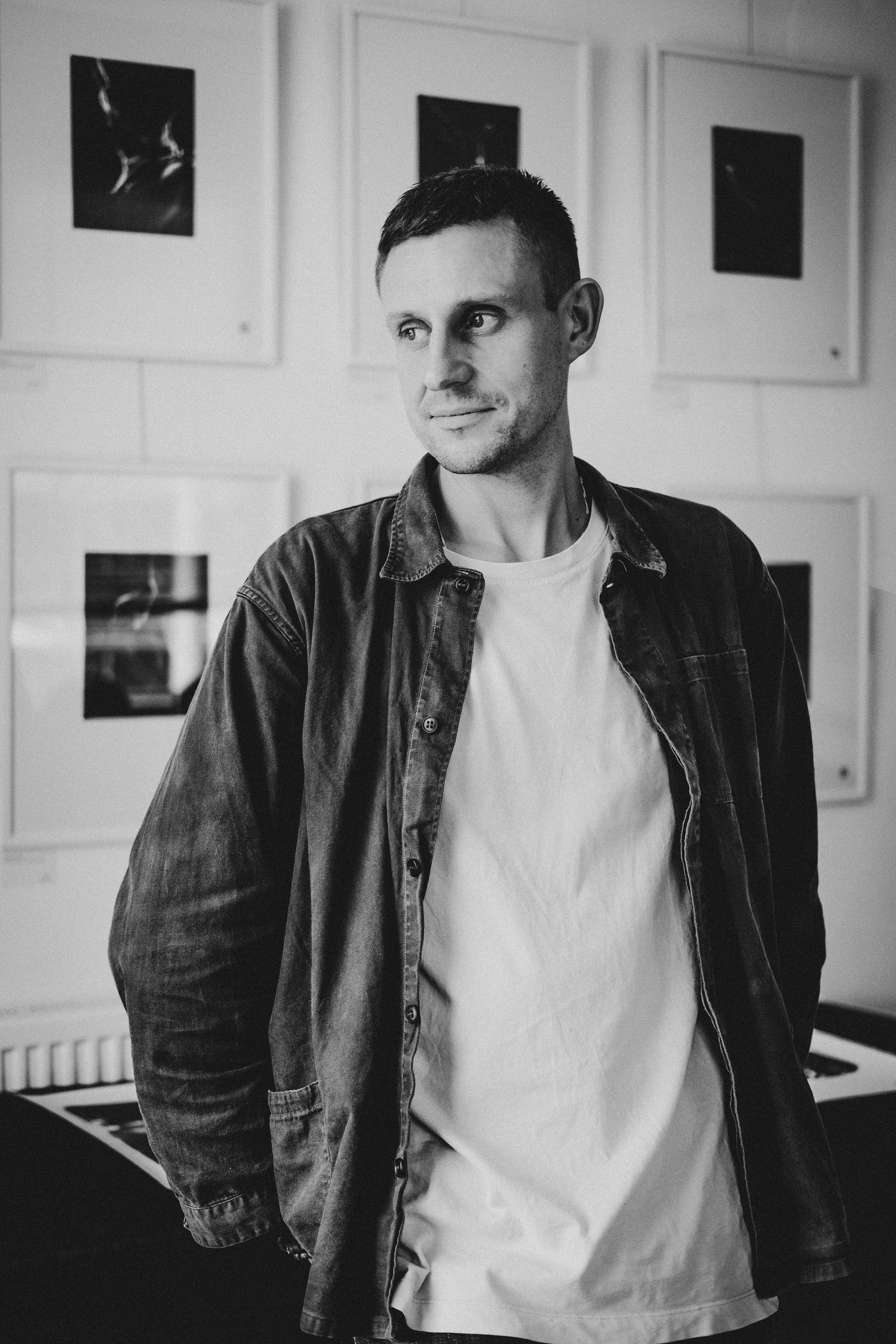 A young man with short hair wearing a white t-shirt and a dark denim jacket standing indoors against a wall with framed pictures.