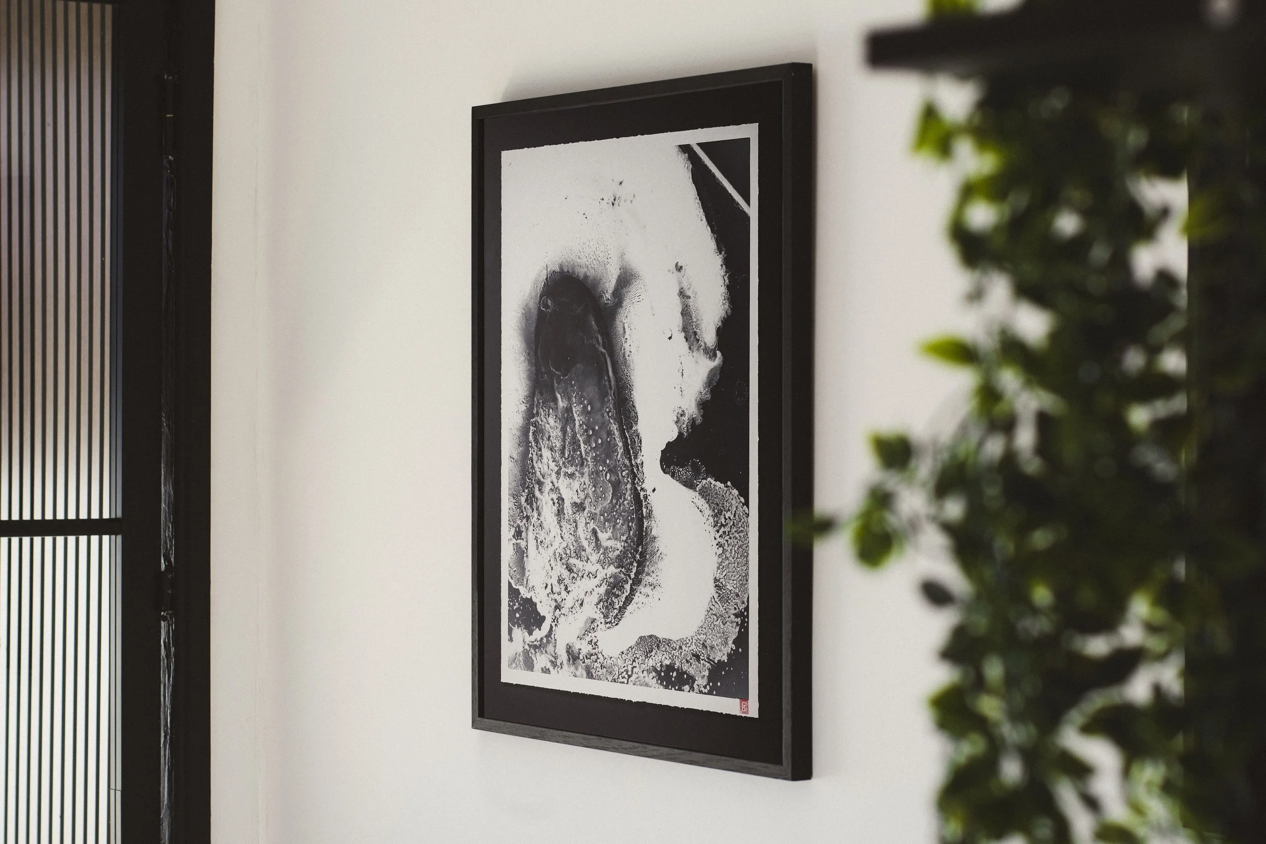 Black and white abstract artwork in a black frame hanging on a white wall, with a green plant partially visible on the right side.