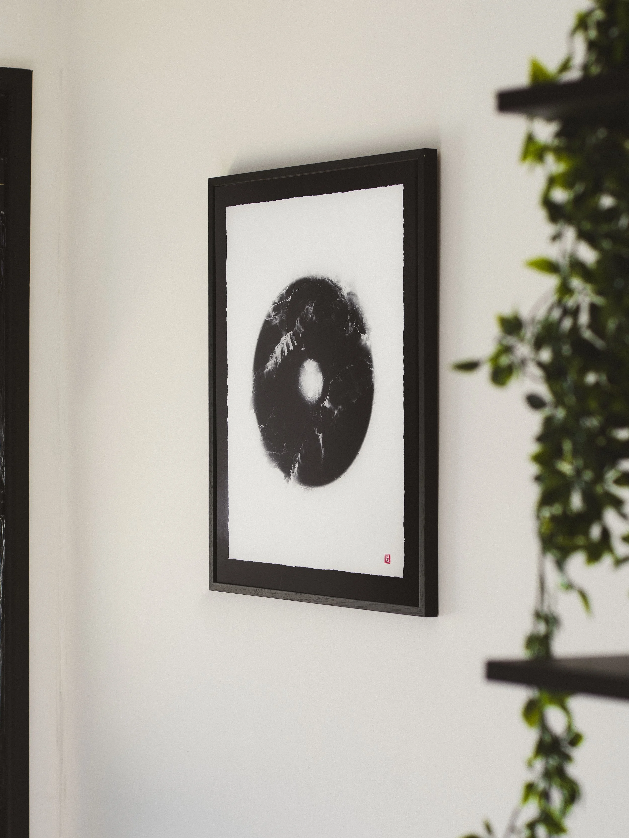 Black and white abstract artwork framed on a white wall, with a small red stamp in the lower right corner.