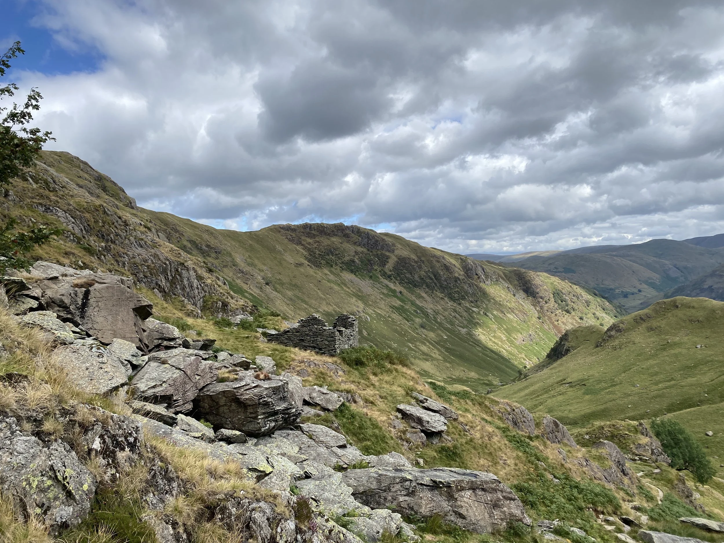 A mountainous landscape above Brotherswater, Lake District UK, with rocky slopes, green grassy hills, and a cloudy sky overhead.