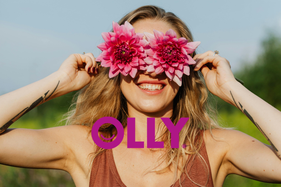 A woman with blonde wavy hair smiling, holding pink flowers over her eyes, wearing a sleeveless brown top, outdoors on a sunny day.