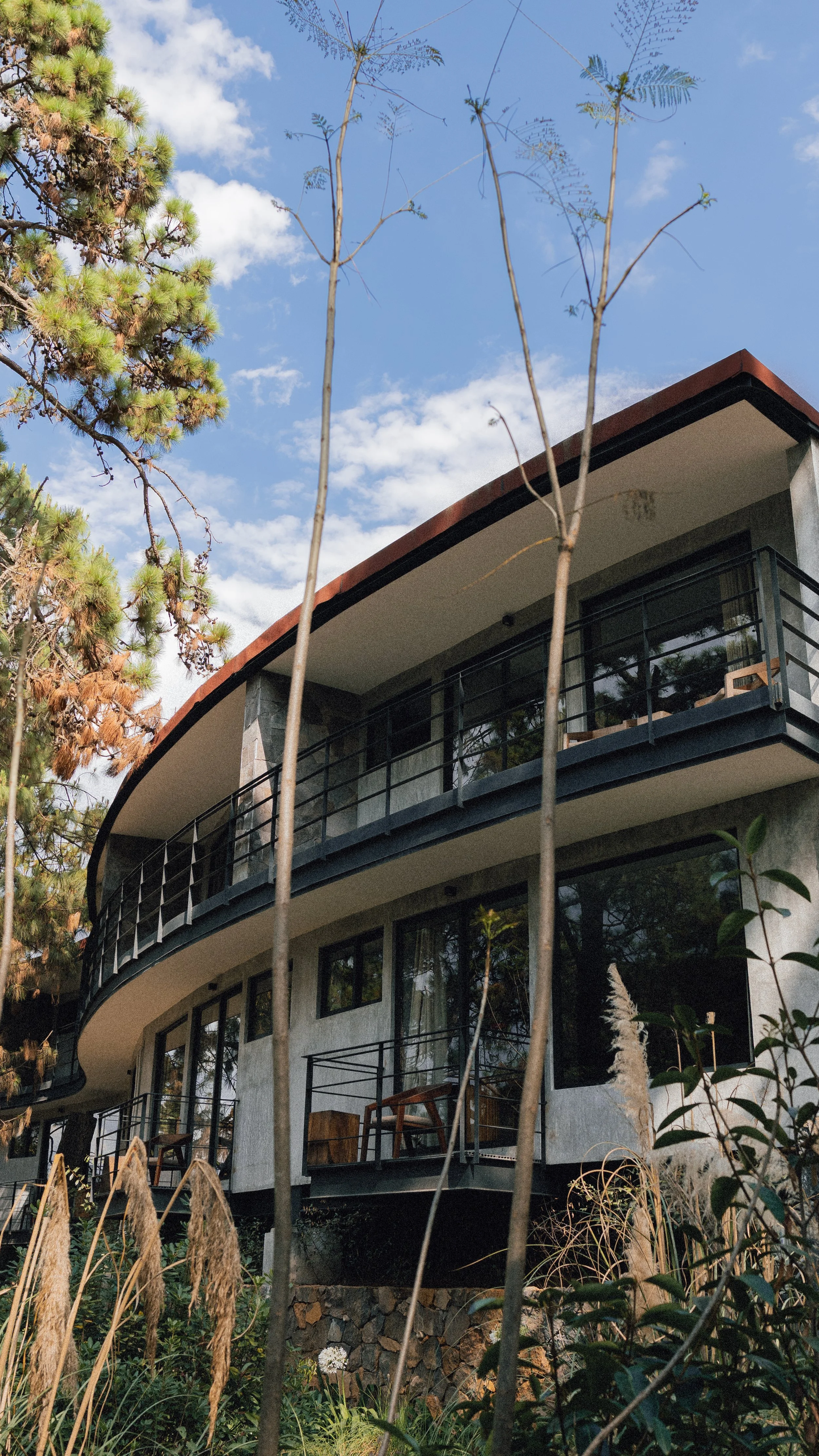 Vista de una casa moderna de múltiples pisos con balcones de vidrio y barandas negras, rodeada de vegetación y árboles, con cielo con nubes.