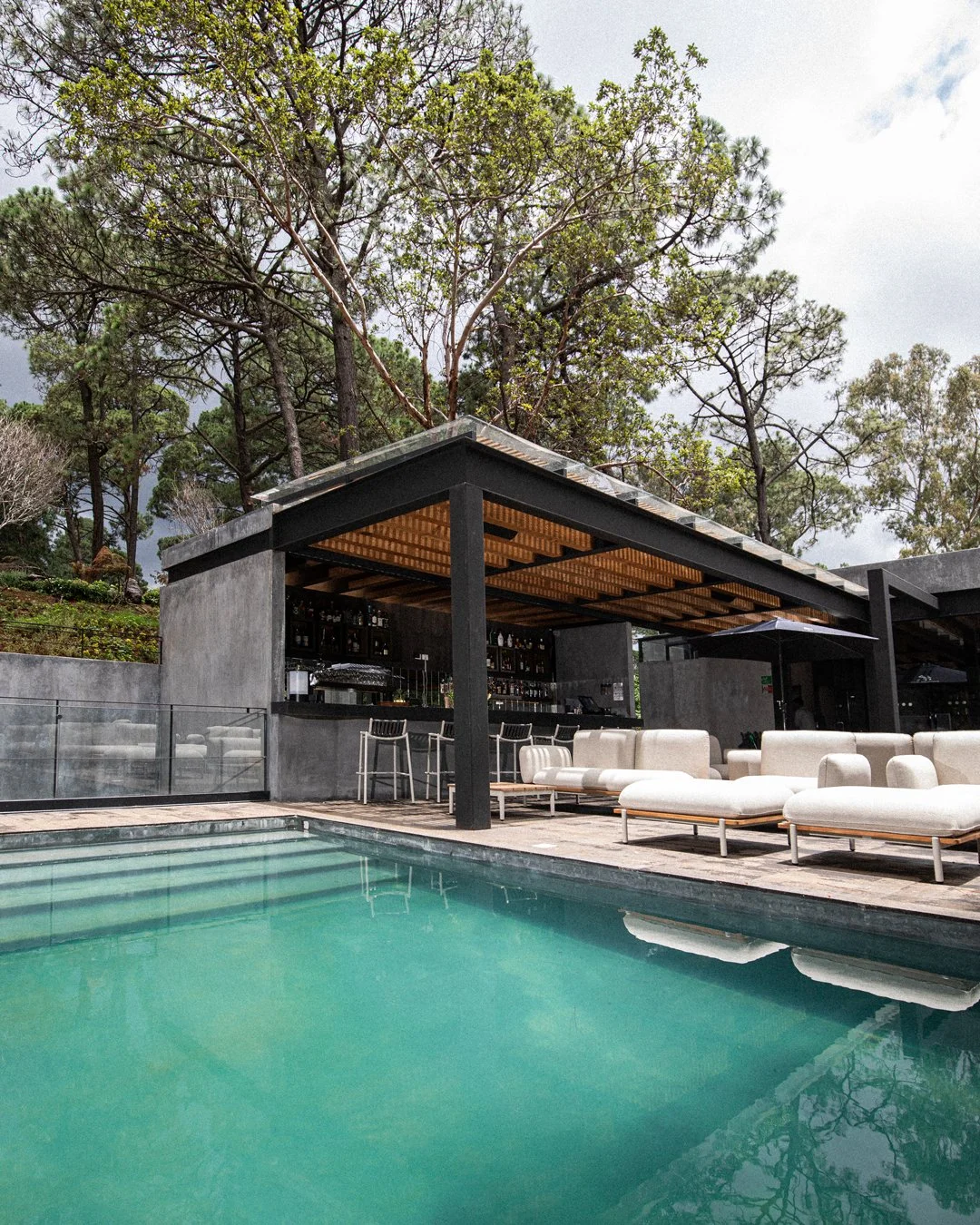 Área de alberca con muebles blancos y una barra de bar techada, rodeada de árboles y cielo nublado.