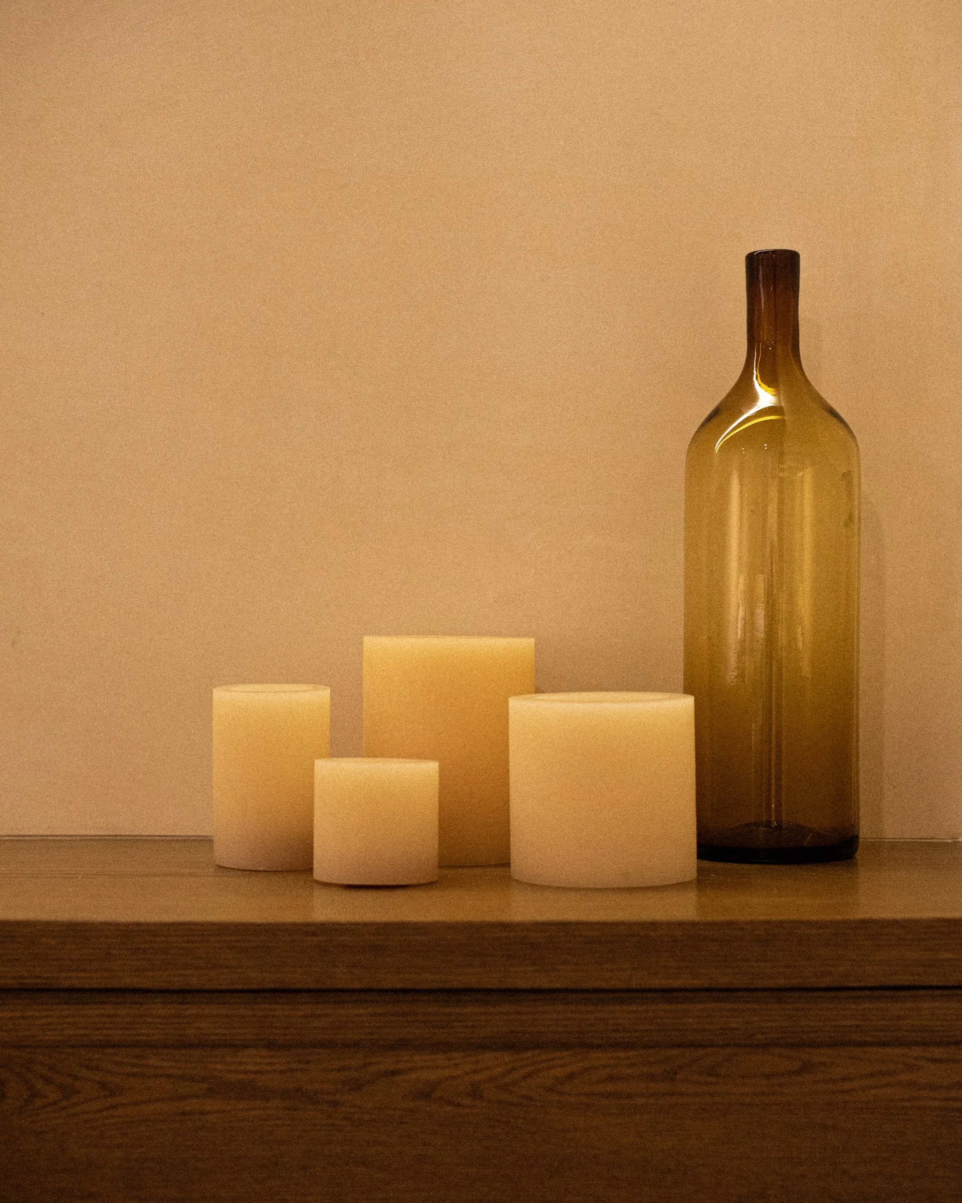 Cuatro velas blancas y una botella de vidrio ambar en una superficie de madera, fondo beige.