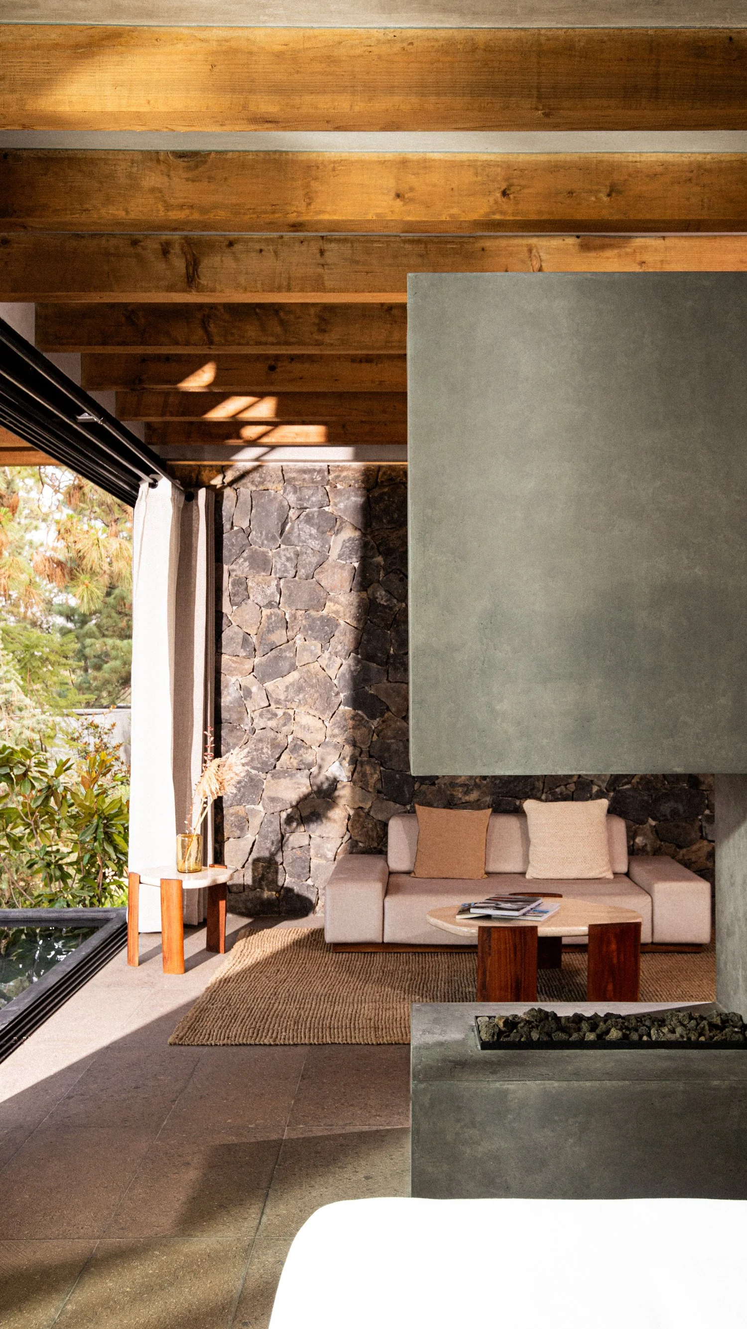 Sala de estar con paredes de piedra, sofá beige con cojines, mesa de café y vista a un jardín con árboles y cortinas blancas, con techo de vigas de madera.