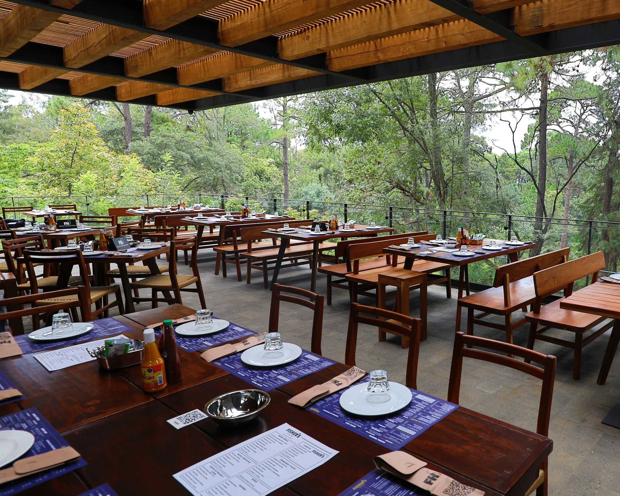 Mesas y sillas de madera en una terraza de restaurante rodeada de árboles verdes