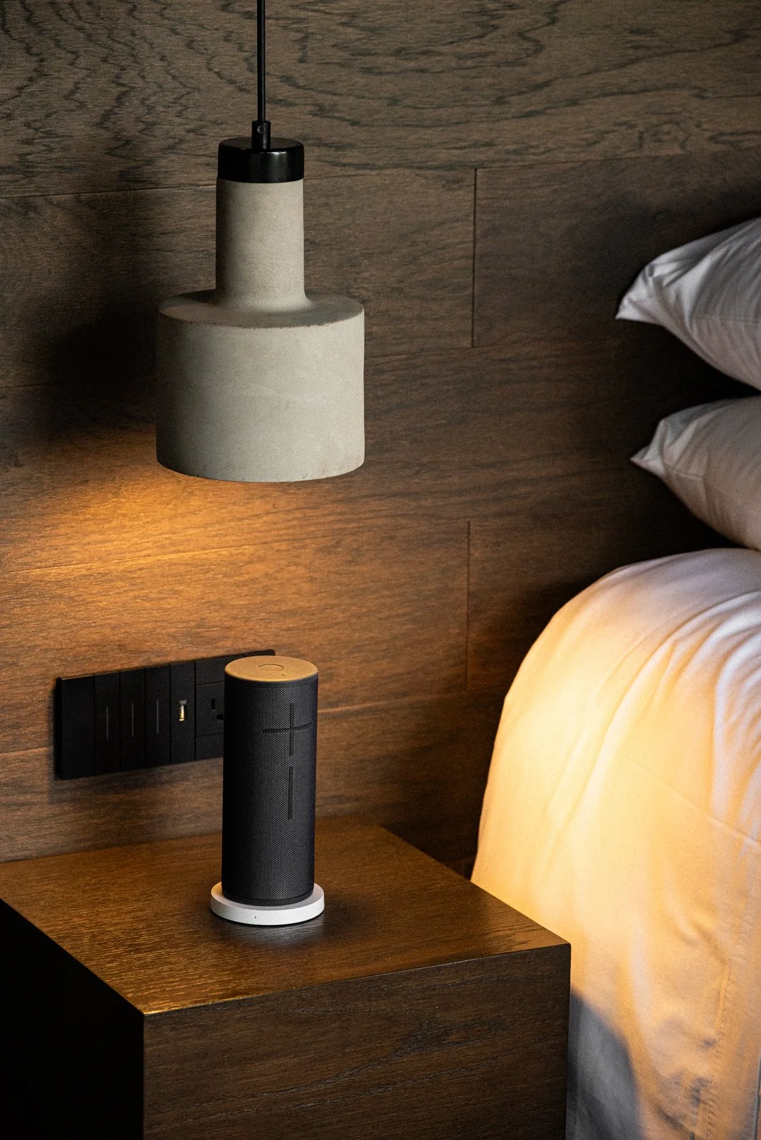 Lámpara de pared, altavoz en la mesita de noche, cama con sábanas blancas, pared de madera.