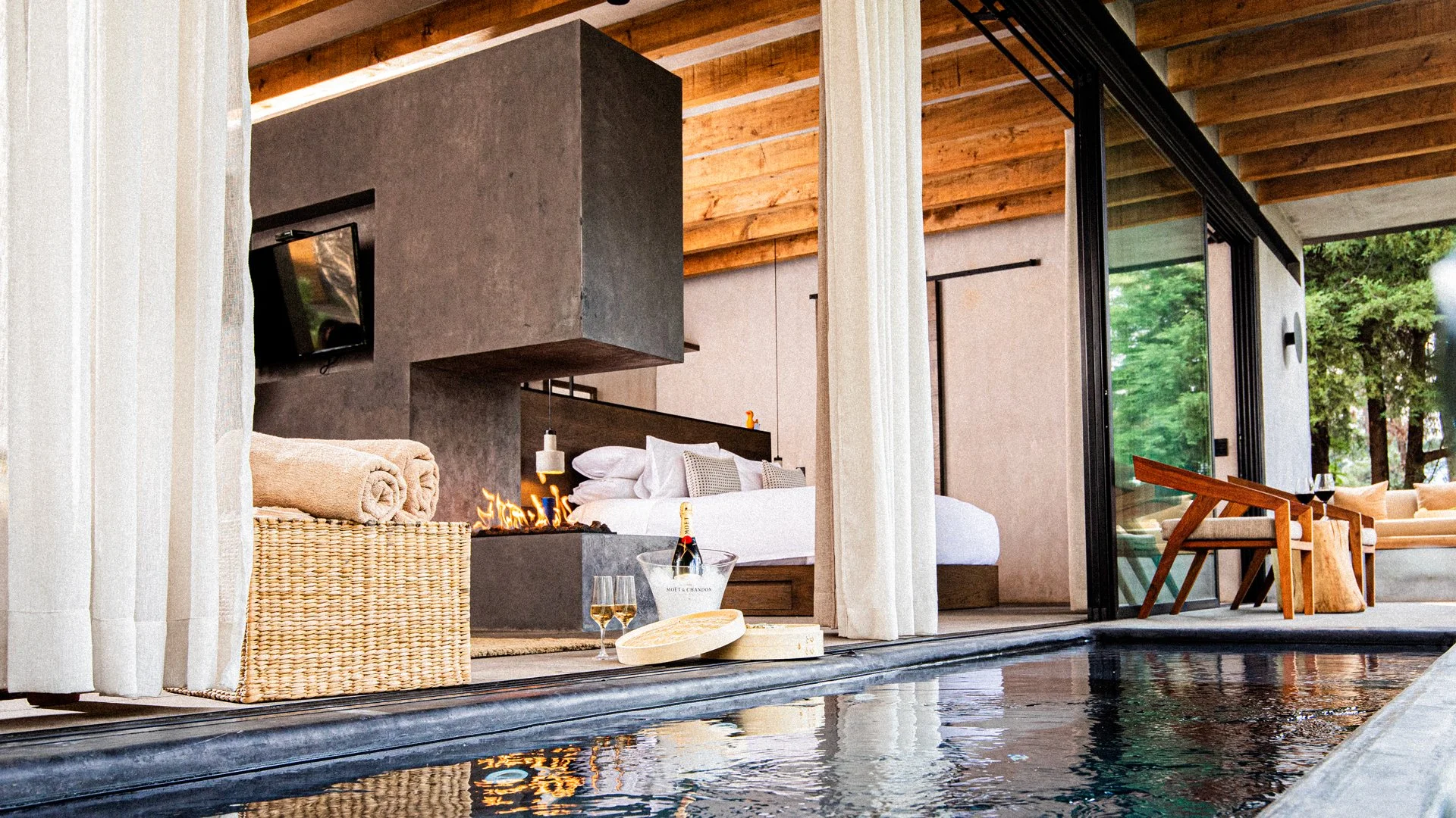 Interior de una habitación moderna con puerta de vidrio corrediza que da a un área exterior con sillones y mesas, vista a árboles verdes, con un área de piscina en primer plano y una decoración elegante que incluye un vino espumoso y un hielo en una cubeta.