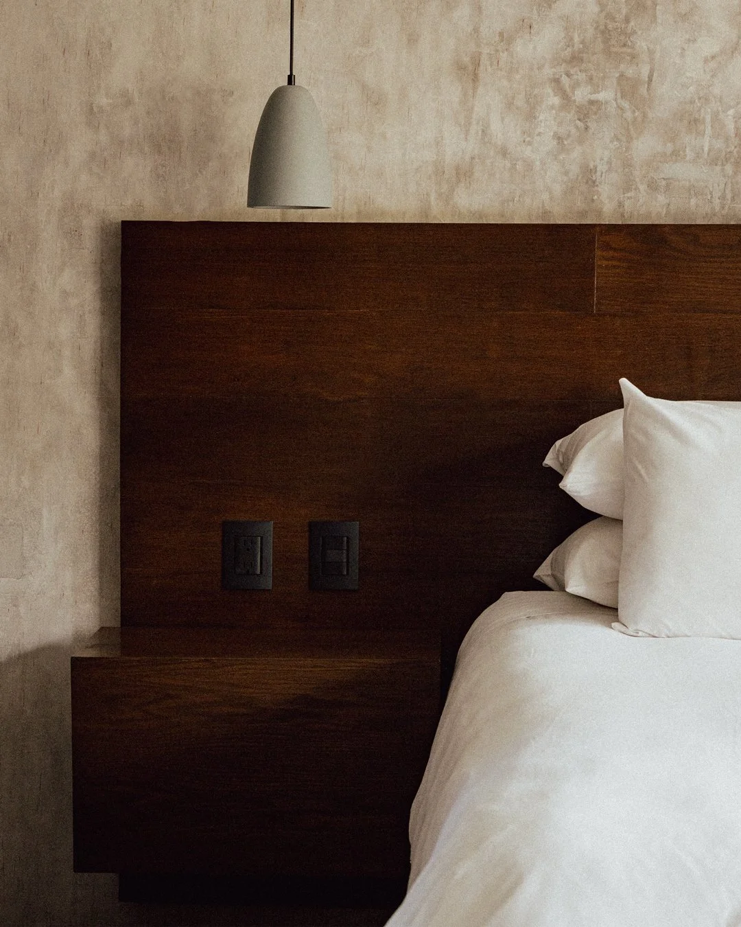 Encabezado de cama de madera oscura con lámpara de pared y paredes de color beige.