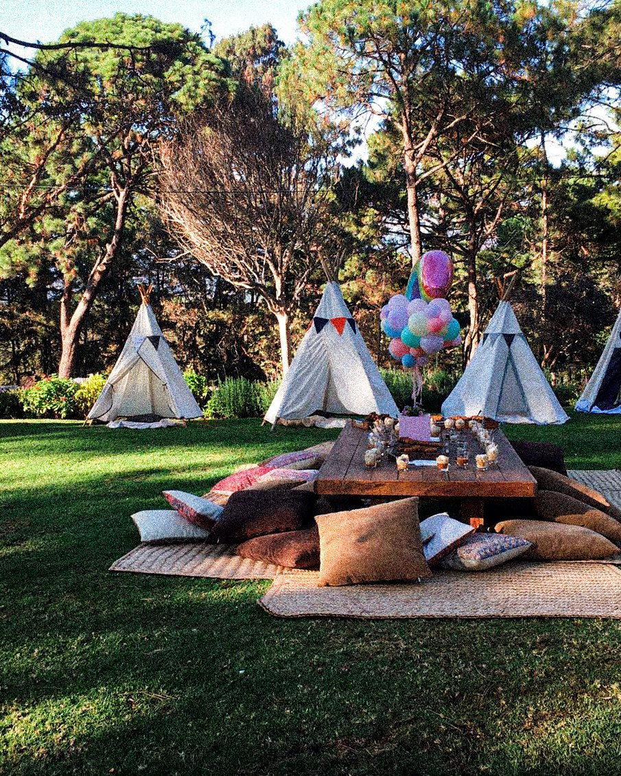 Área de camping al aire libre con tiendas tipi, un círculo de almohadas y una mesa en el centro con decoraciones, globos de colores y árboles de fondo.