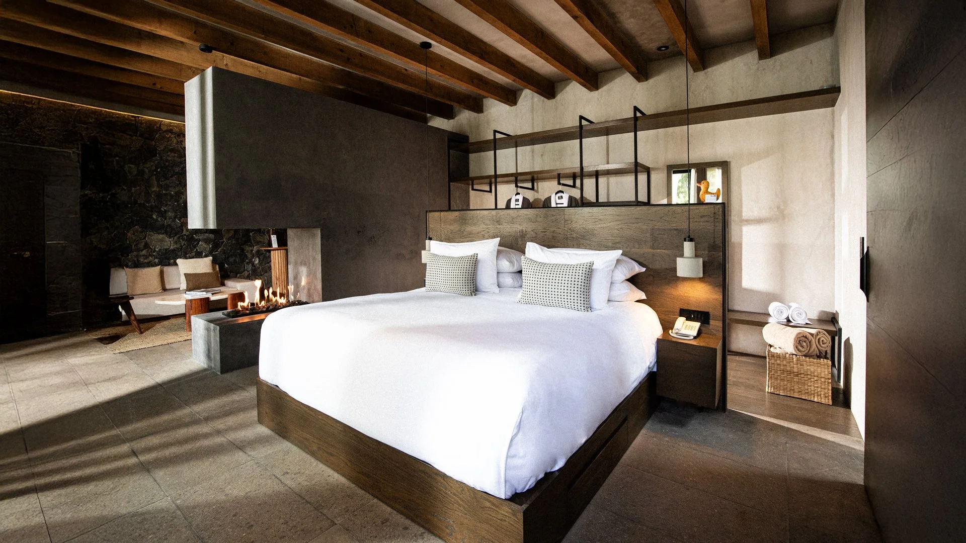 Hotel moderno con cama grande, paredes de concreto y decoración de madera, con un área de sillones, chimenea y accesorios en tonos neutros.
