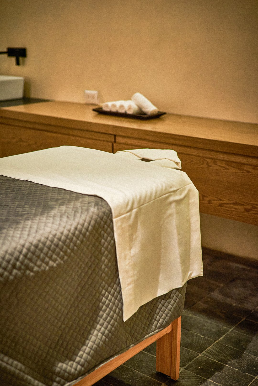 Cama en una habitación con sábanas y una mesa de madera en el fondo con toallas enrolladas