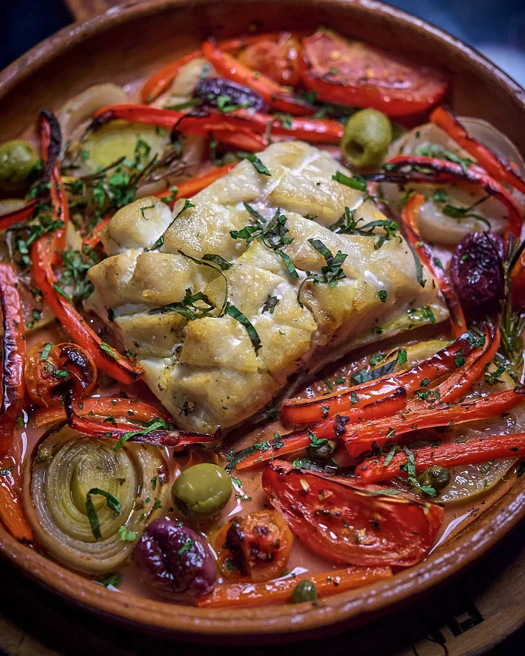 Pescado horneado en una cazuela con pimientos rojos, cebolla, aceitunas verdes y hierbas frescas.