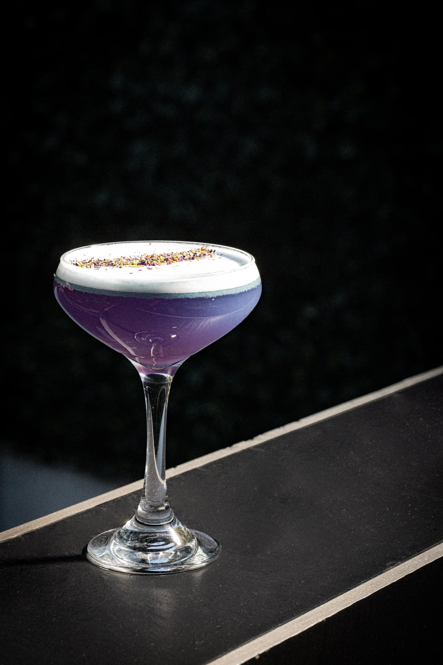 Copa de cóctel con bebida violeta y espuma blanca en la parte superior, decorada con pequeñas escamas de color en la parte superior, colocada sobre una superficie negra.